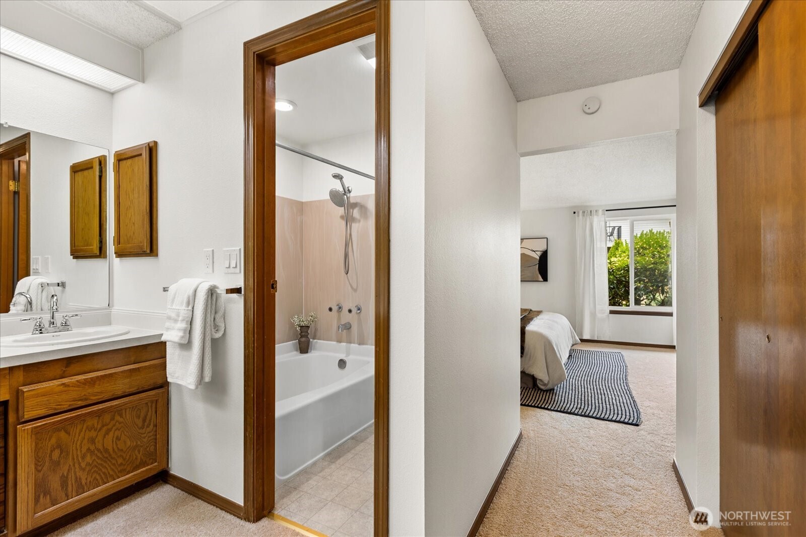 Bathroom vanity and tub beside a hallway opening onto a bedroom with a striped rug.