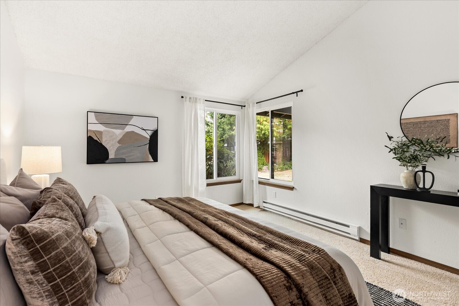 Bedroom with a large cream-and-brown dressed bed beside two curtained windows overlooking greenery.