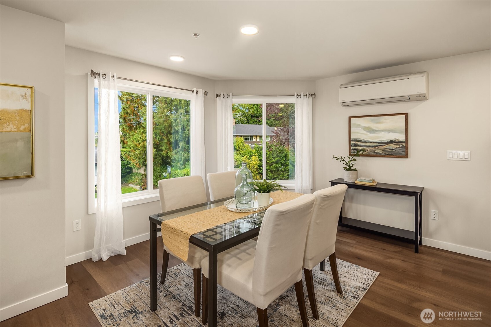 Dining area with a glass-top table, cream chairs, and large windows overlooking greenery.