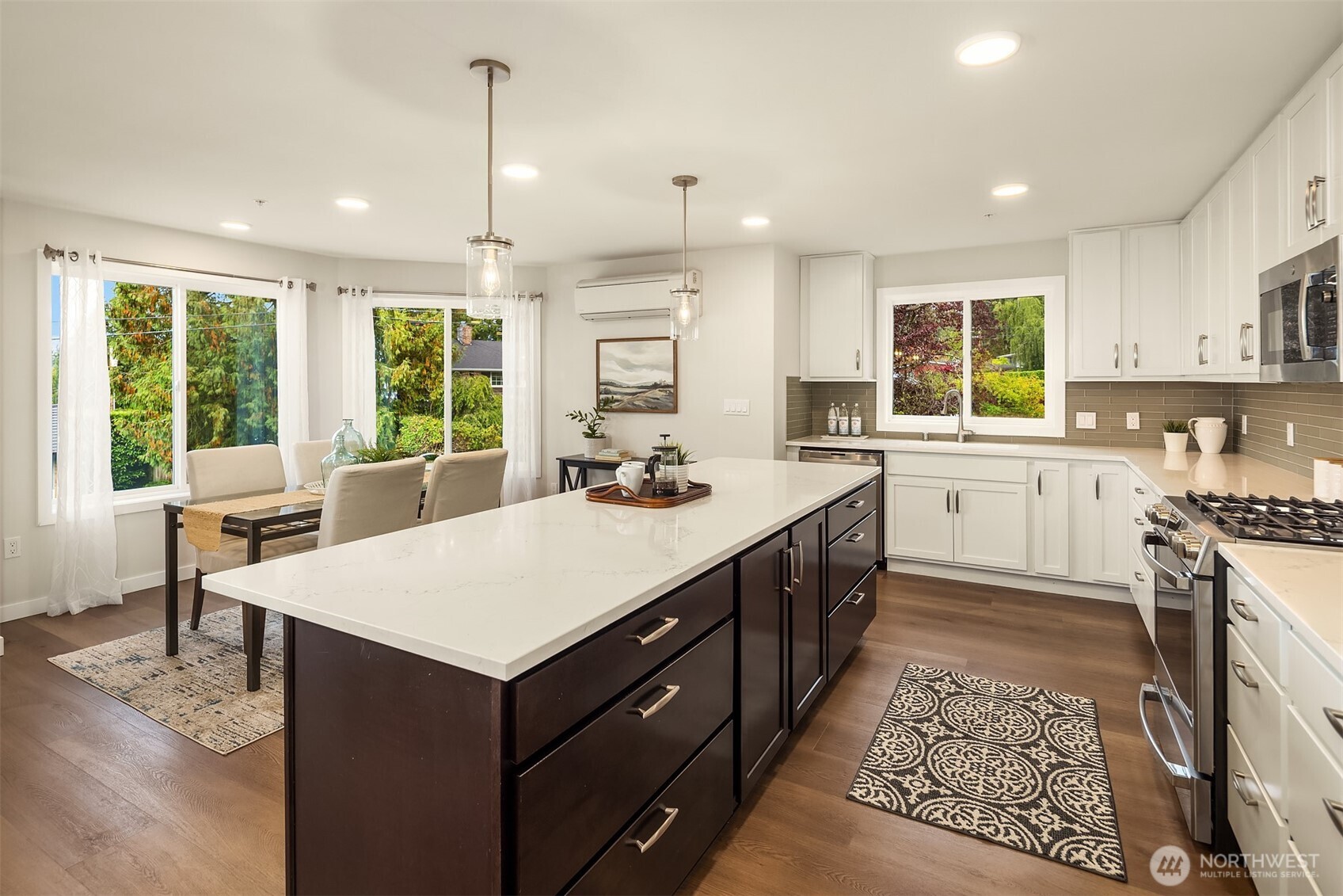Open kitchen and dining area with a large white-stone island, white cabinetry, and windows overlooking greenery.