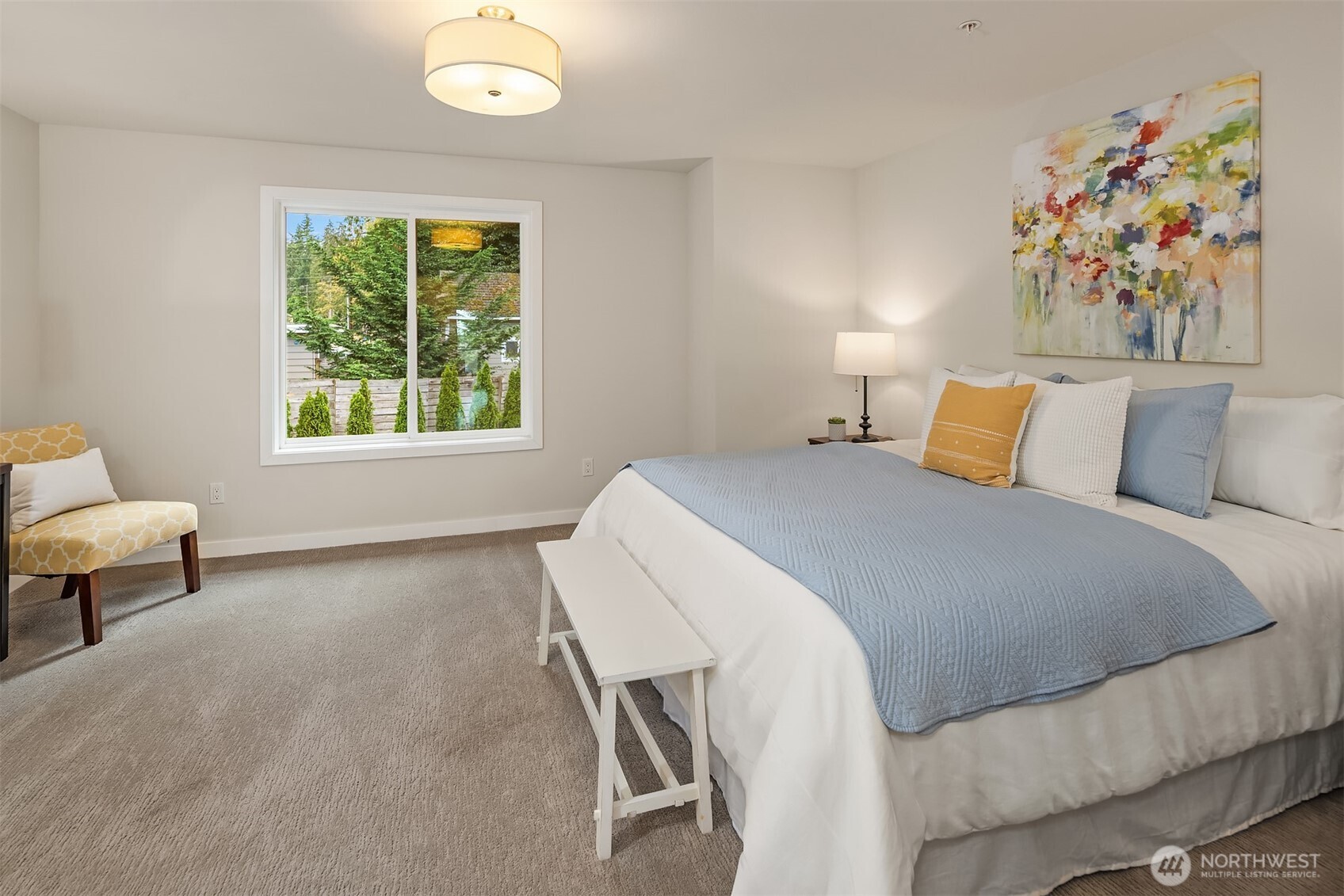 Staged bedroom with a large bed, pale blue quilt, abstract wall art, and a window overlooking trees.