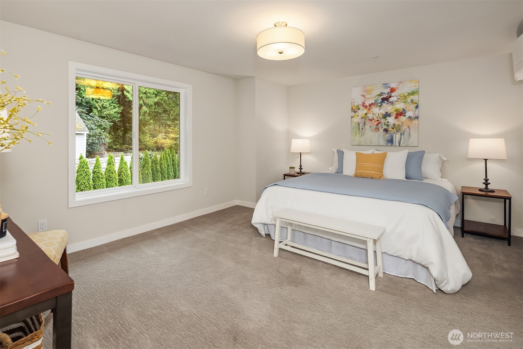 Staged bedroom with a white and blue bed beside a wide window overlooking evergreens.
