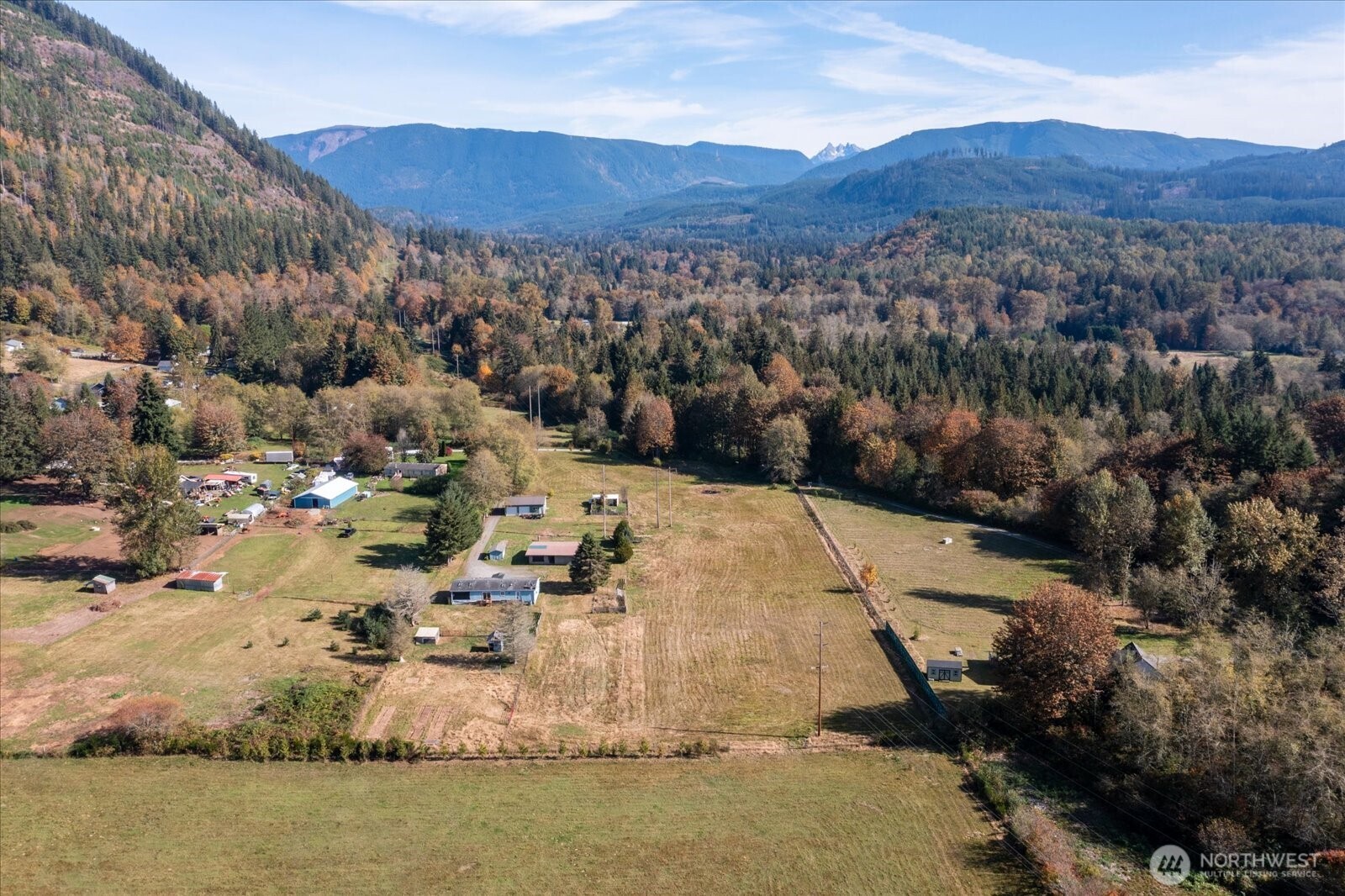 Aerial view of a rural property with open fields, outbuildings, and forested mountain views.