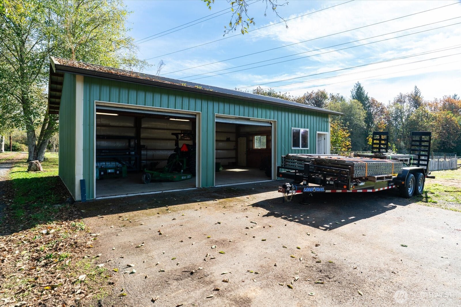 Green metal workshop with two open garage bays and a loaded flatbed trailer in front.