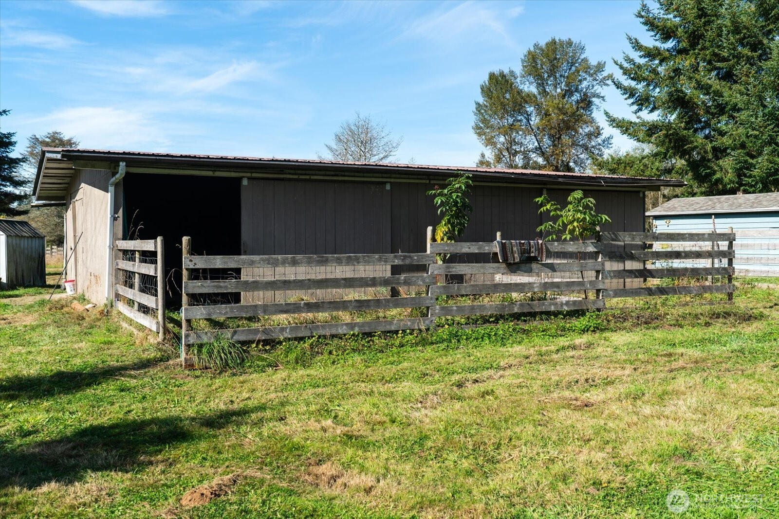 Dark-brown barn with an open bay behind a wooden fenced enclosure.