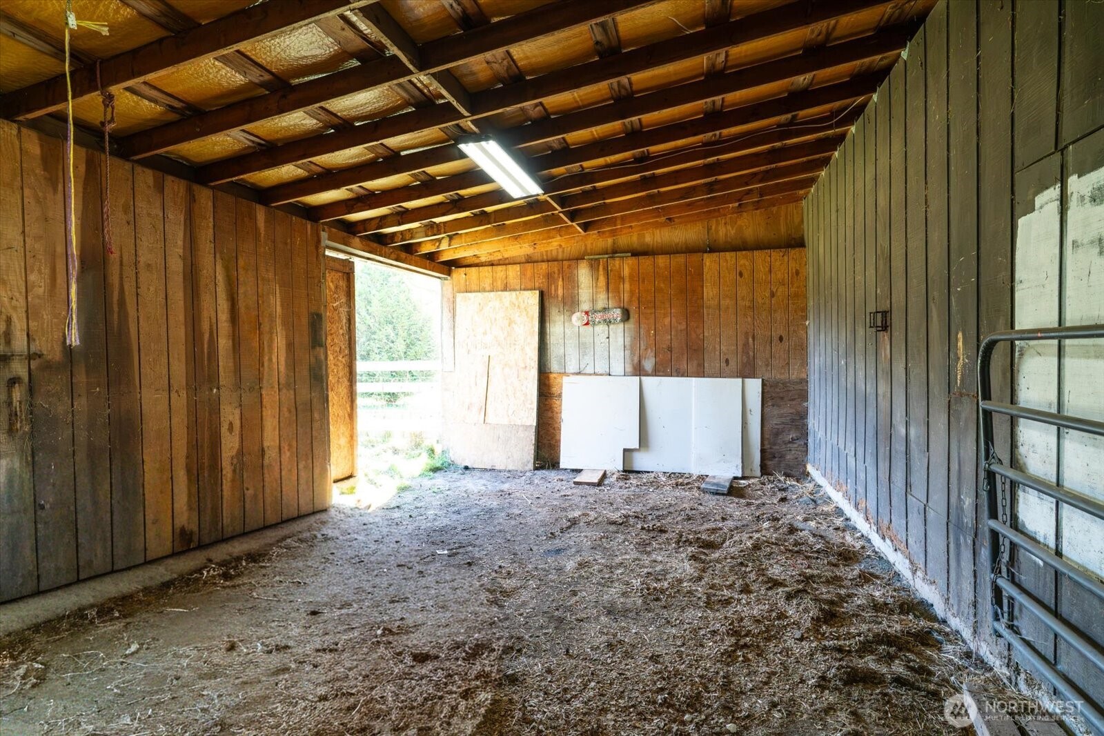 Rustic barn interior with exposed rafters, a dirt-and-straw floor, and a metal stall gate.