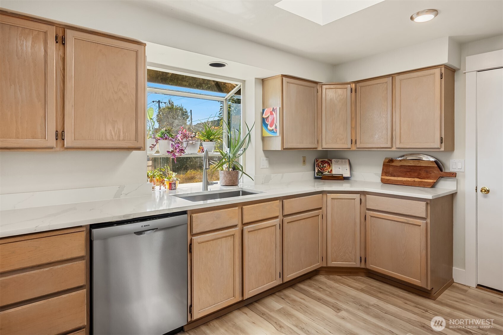 Kitchen with light wood cabinets, white counters, and a plant-filled window above the sink.