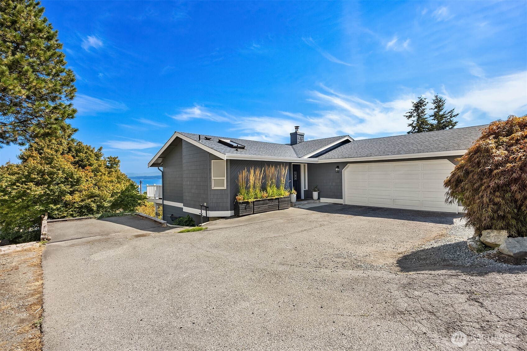 Gray house with a white double garage, wide driveway, and water visible through the trees.
