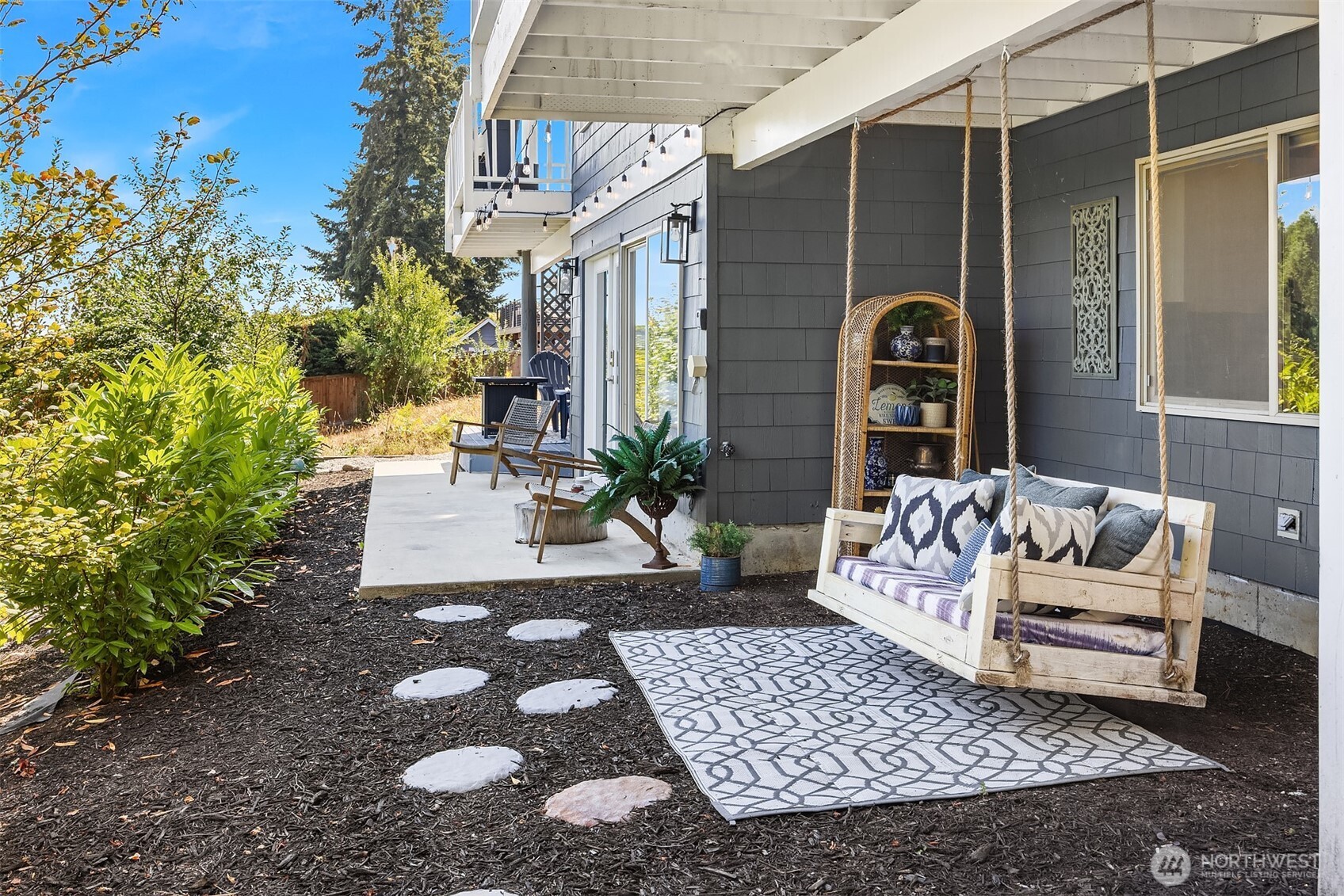 Covered patio with a cushioned hanging swing, patterned rug, and stepping-stone path beside the house.