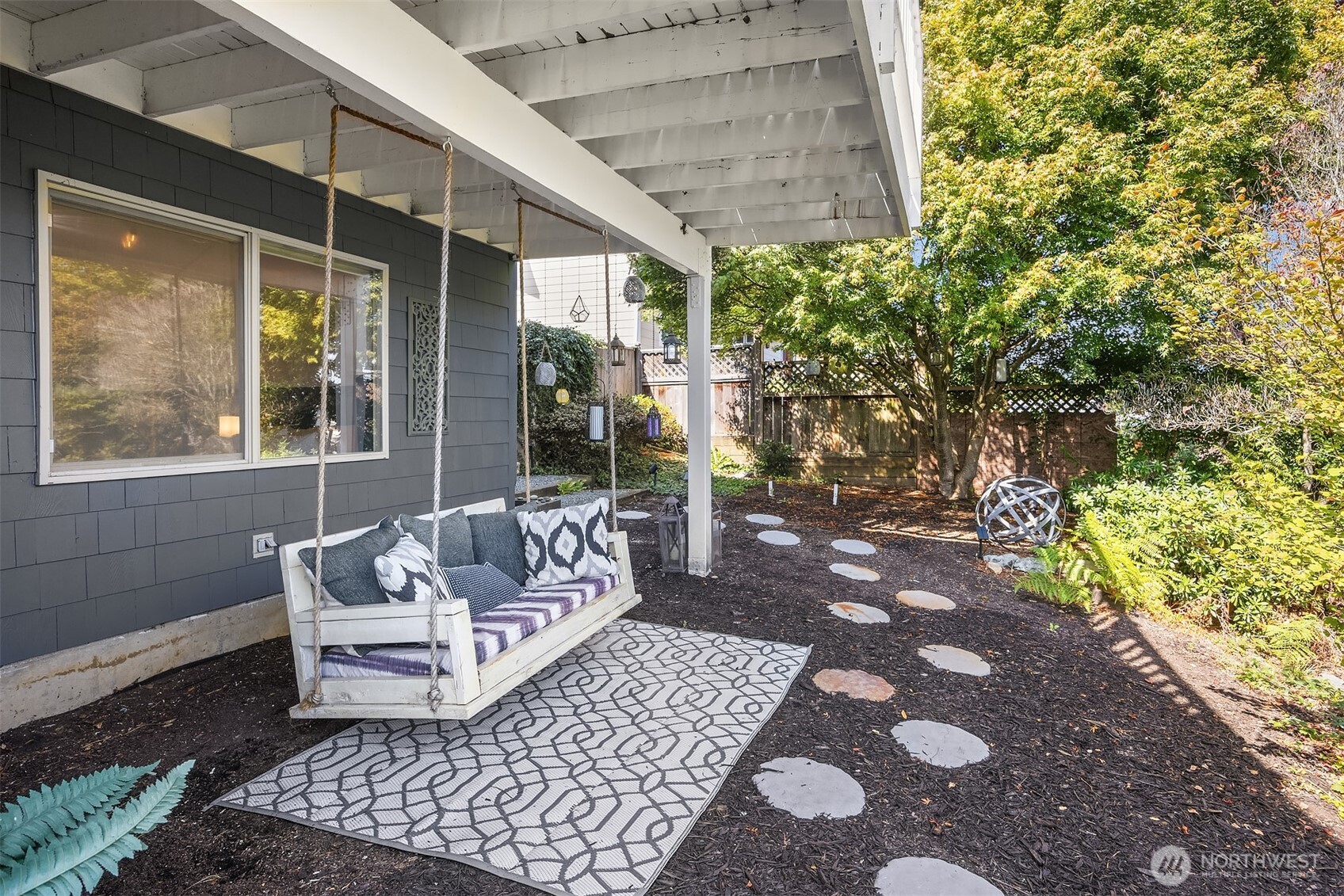 Covered patio with a cushioned hanging swing, patterned rug, and stepping-stone path through a leafy backyard.