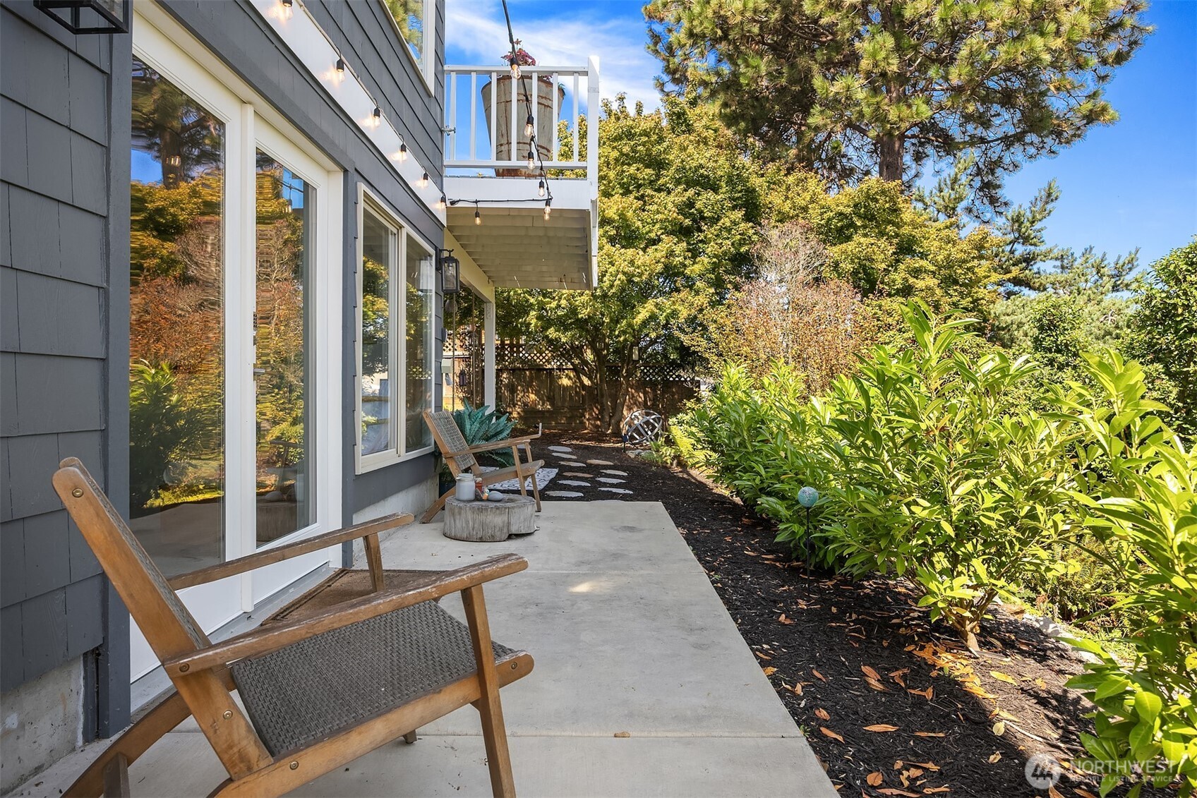 Home’s side patio with sliding glass doors, wooden chairs, and a garden bordered by mature trees.
