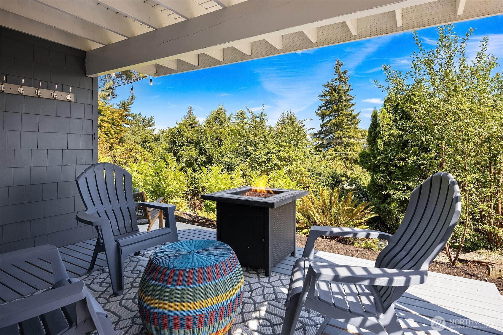 Covered deck with Adirondack chairs, a lit fire pit, and lush garden landscaping.