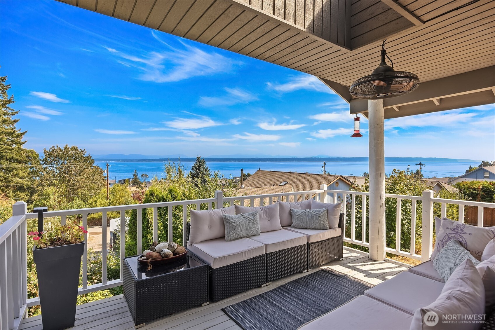 Covered deck with wicker seating overlooking blue water and a distant shoreline.