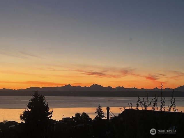 Sunset over calm water and a jagged mountain range, framed by silhouetted trees and rooftops.