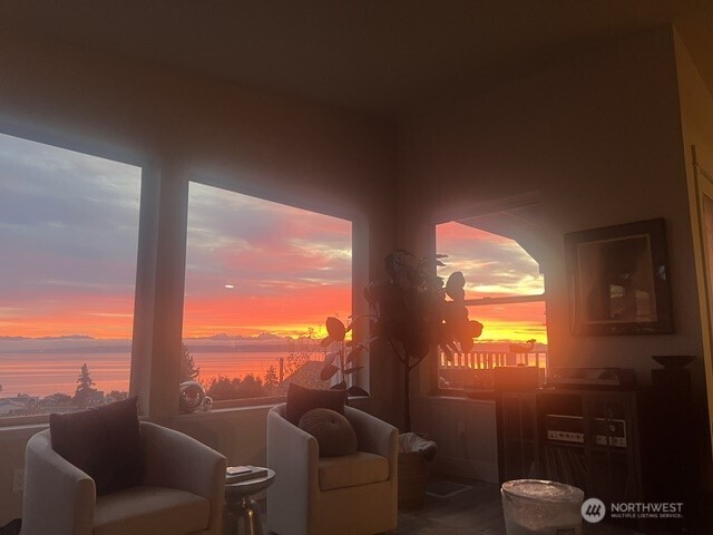 Furnished living room with broad windows overlooking water and distant mountains at sunset.