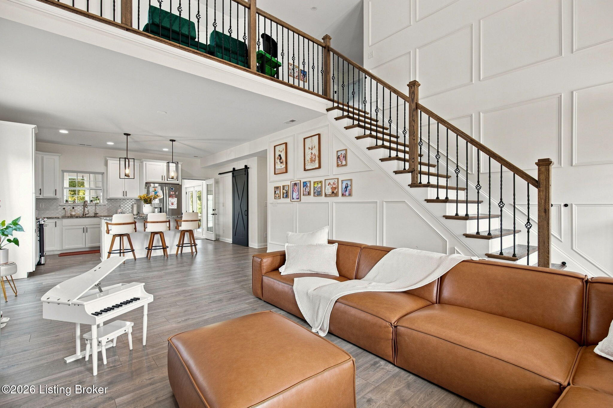 Open living room with a brown leather sectional, white piano, kitchen, and staircase.