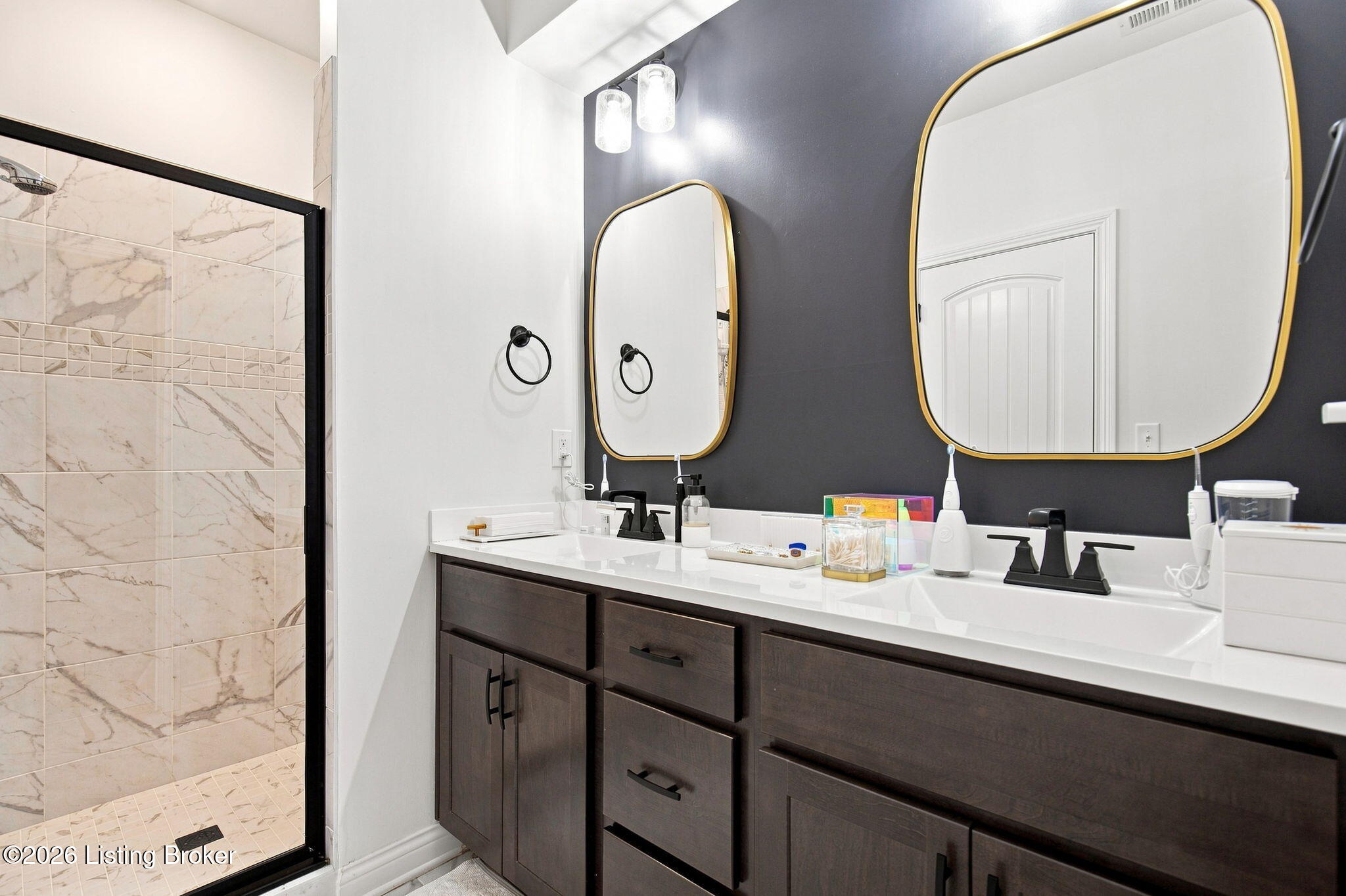 Bathroom with a dark double vanity, gold-framed mirrors, and a marble-look tiled shower.