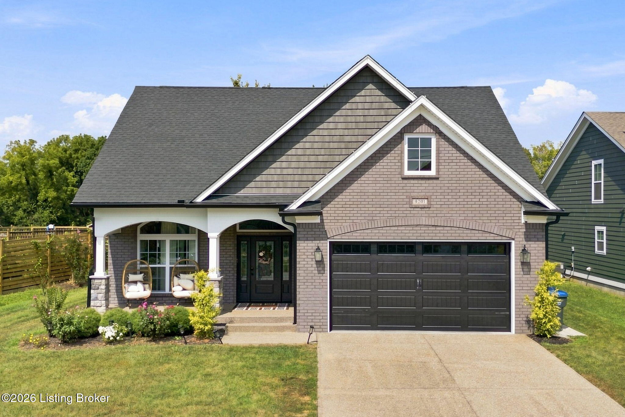 Brick house with a covered porch, hanging chairs, landscaped flower beds, and a double garage.