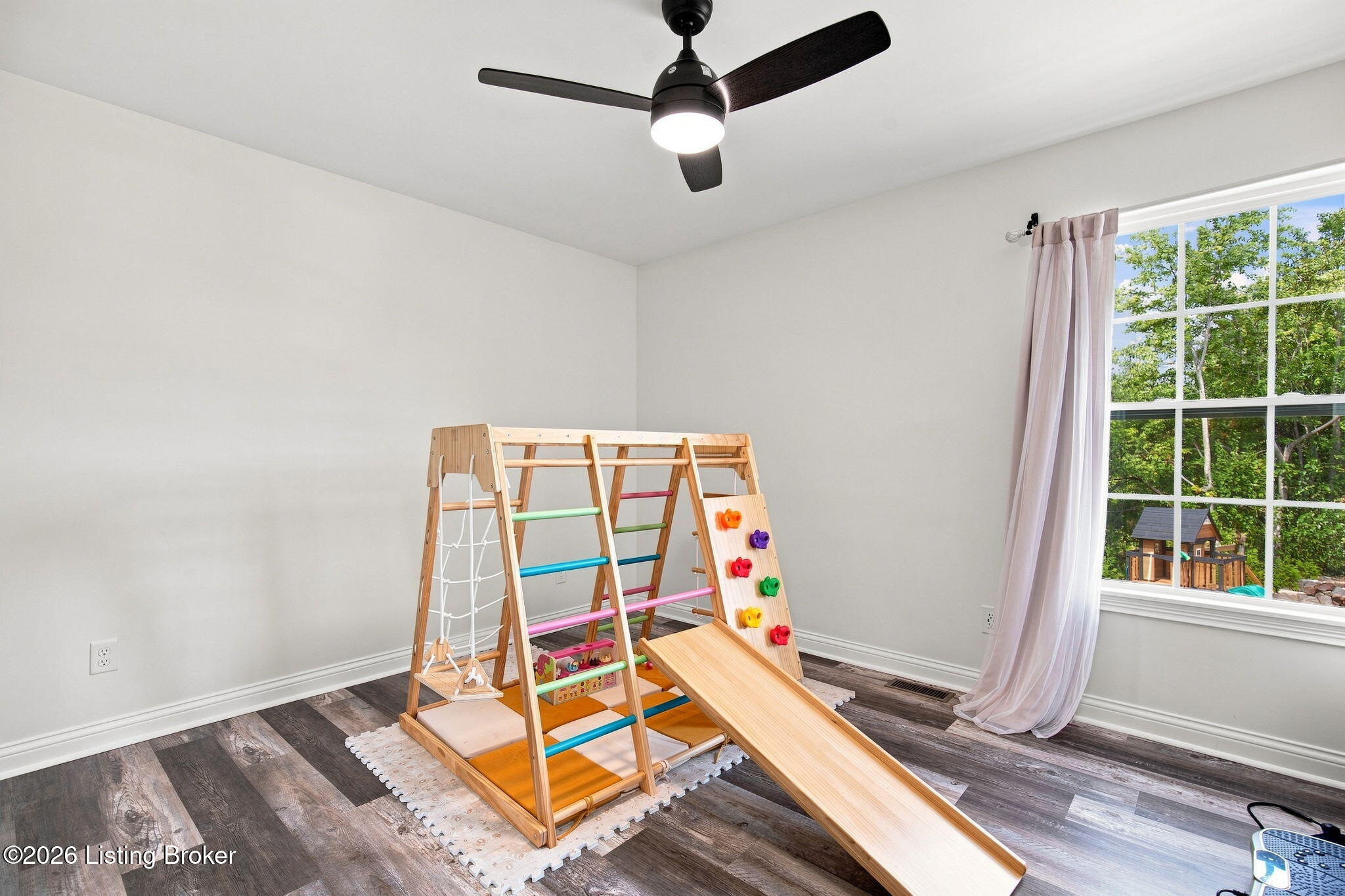 Playroom with a wooden climbing gym and slide beside a large window overlooking trees.