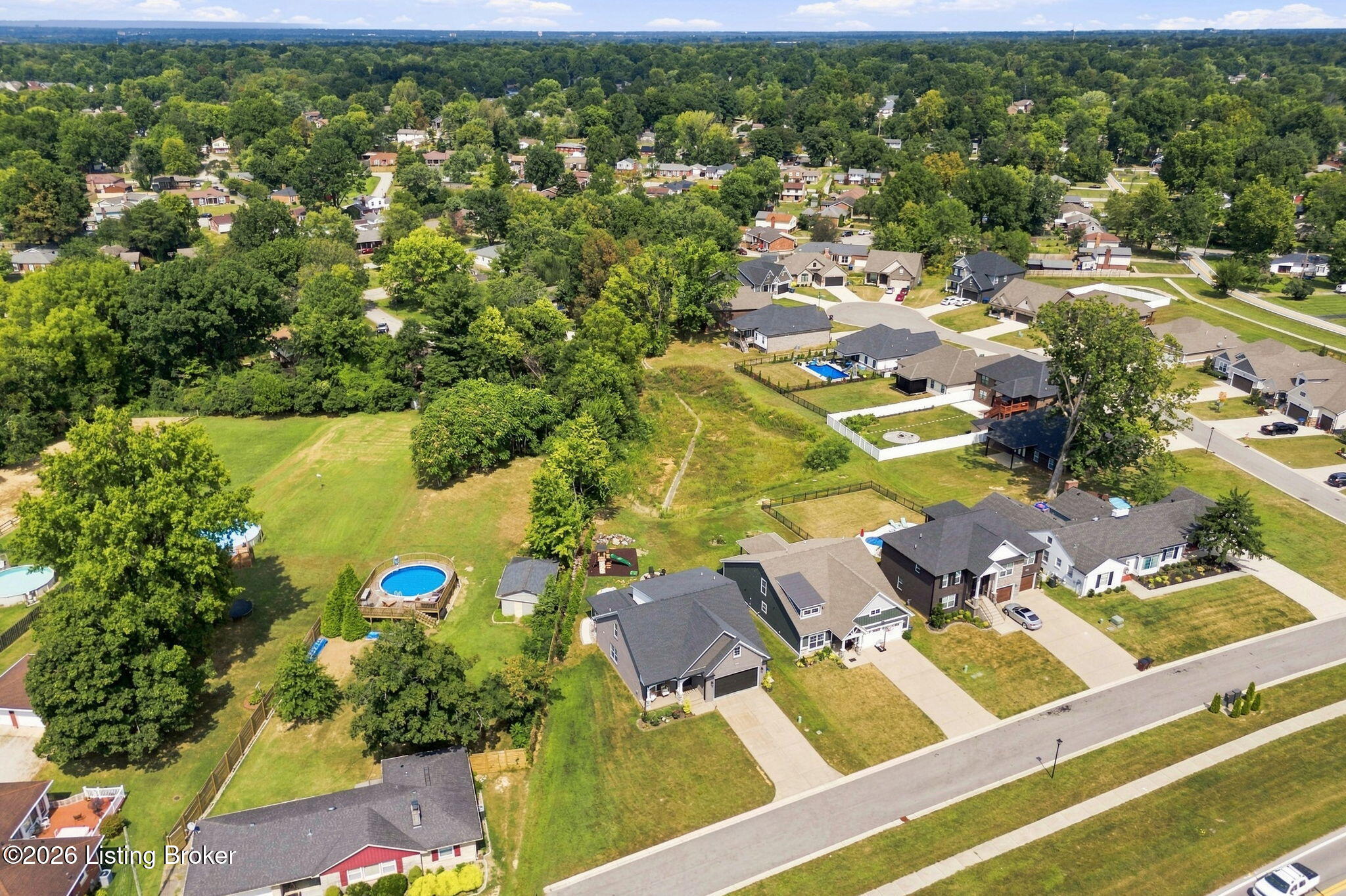 Aerial view of a grassy parcel amid homes, mature trees, and swimming pools.