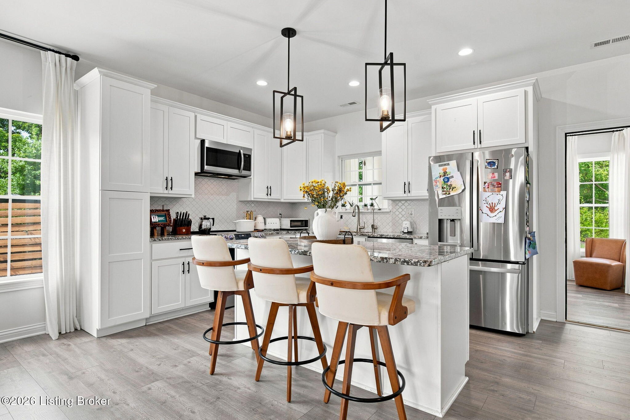 Bright white kitchen with a stone island, three stools, stainless steel appliances, and black pendant lights.
