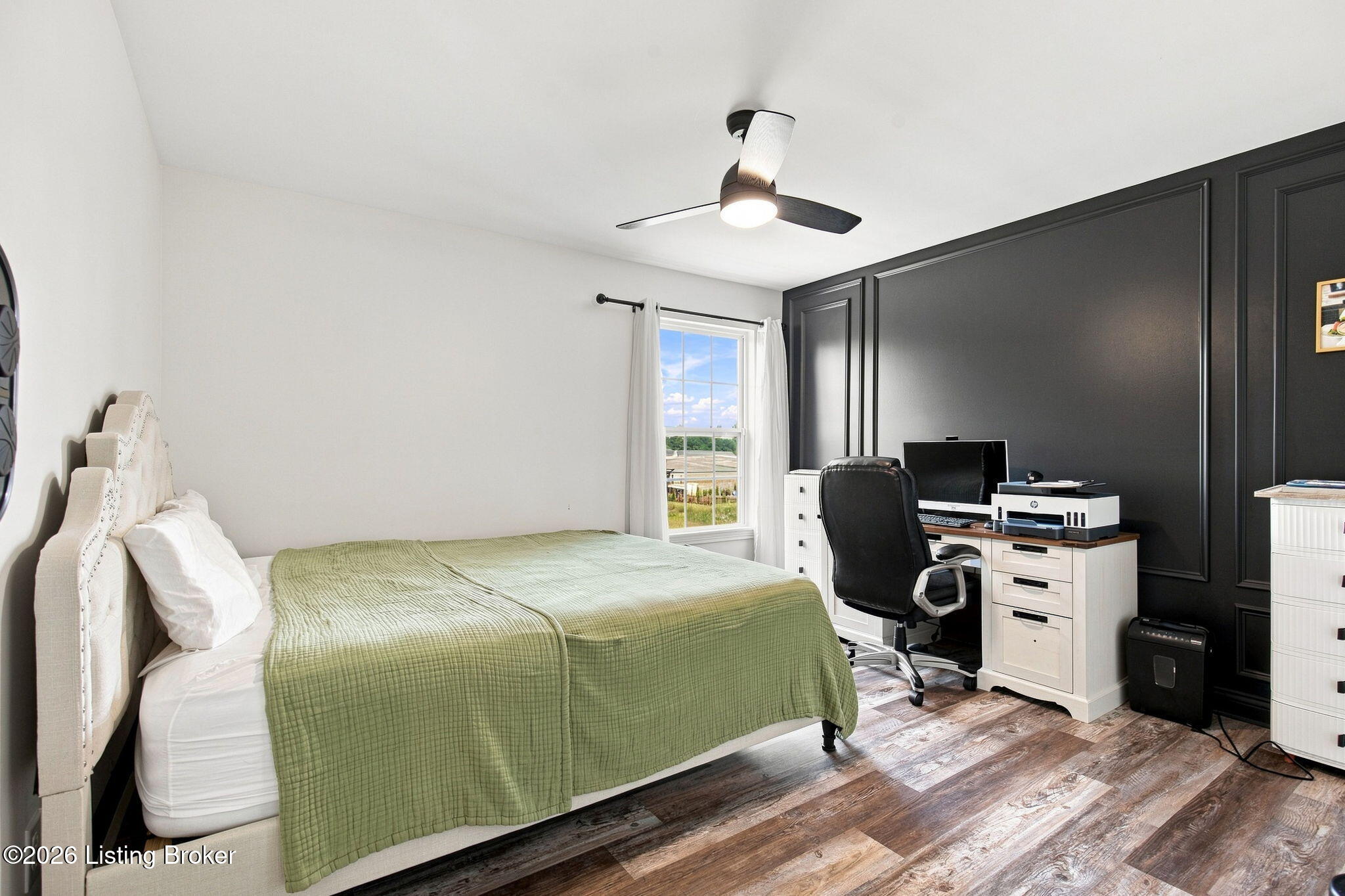Bedroom with a sage-green quilt, dark paneled accent wall, and desk with computer and printer.