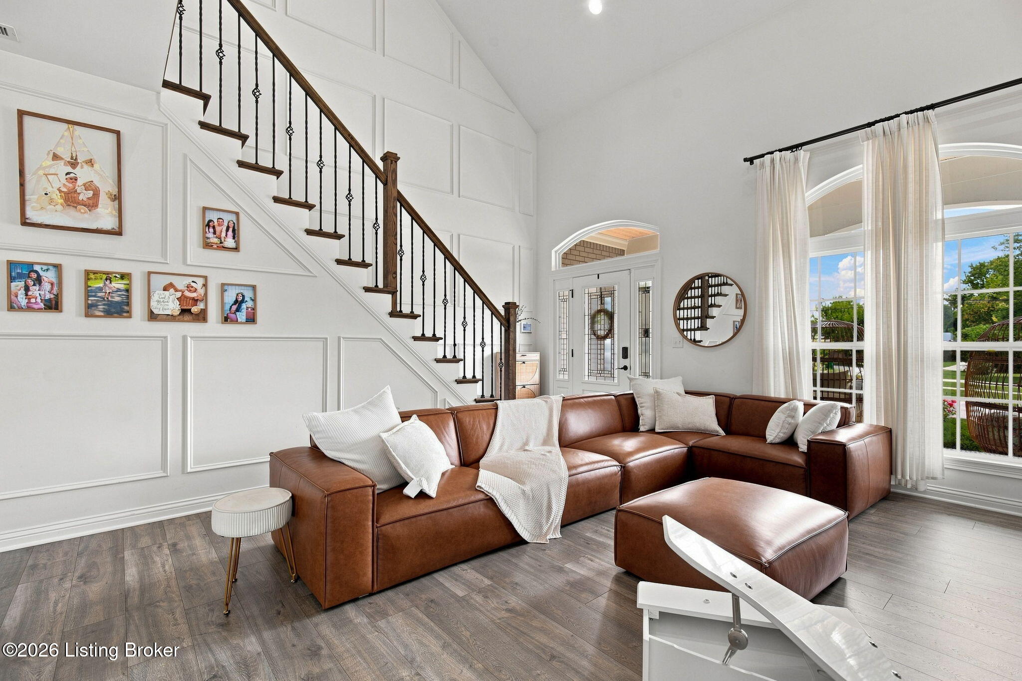 Furnished living room with a brown leather sectional, tall paneled walls, and a dark wood staircase.