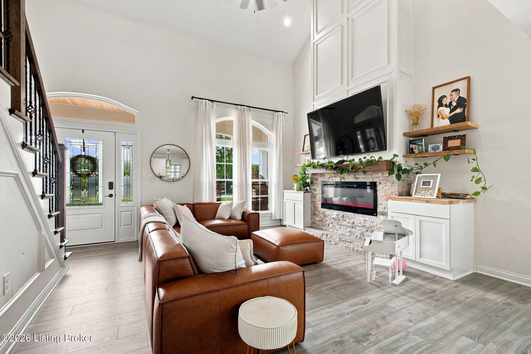 Living room with a brown leather sectional, stone fireplace, tall windows, and a staircase.