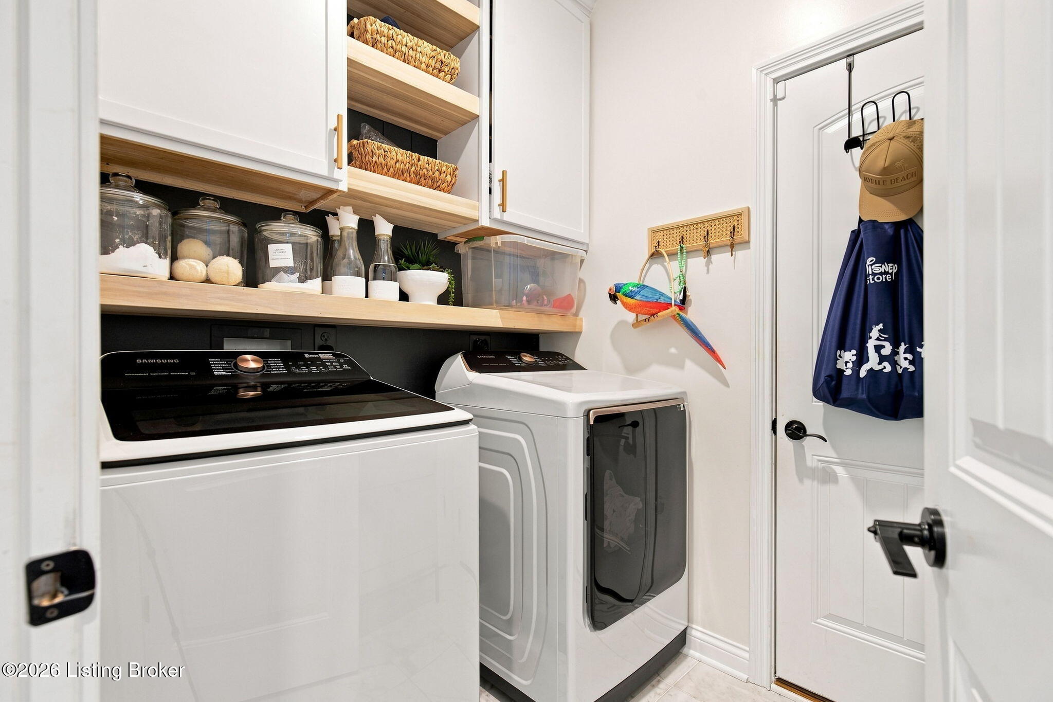 Laundry room with white washer and dryer, open wood shelves, and a colorful parrot wall decoration.