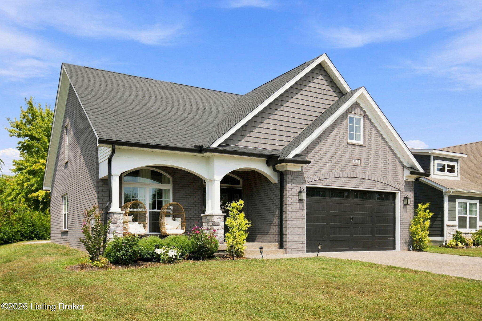 Gray-shingled home with a covered porch, wicker chairs, landscaped beds, and a dark double garage.
