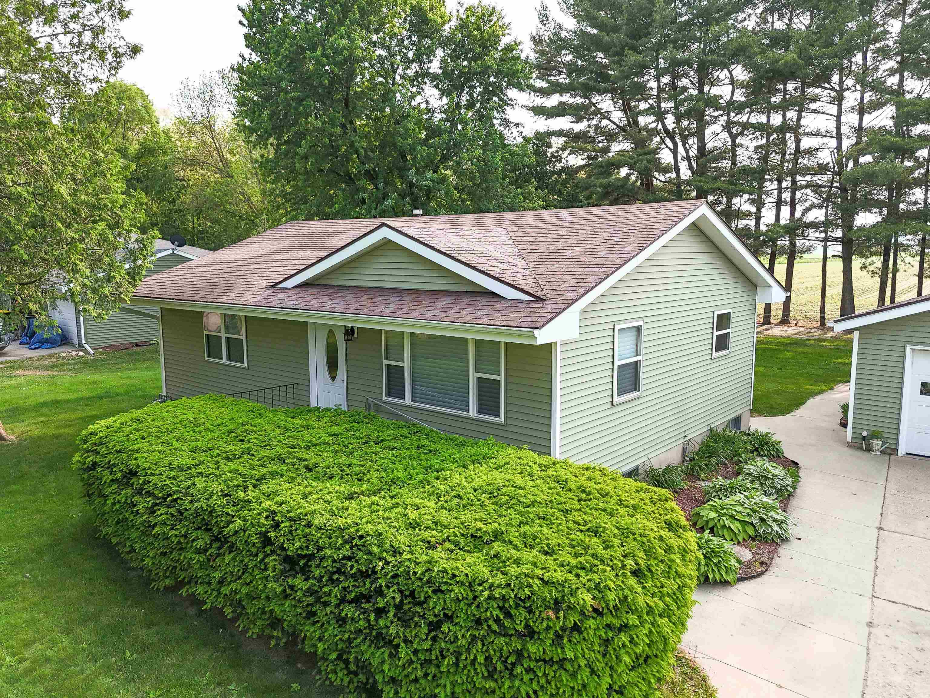 Green single-story house with a brown shingle roof, large front hedge, and concrete driveway.