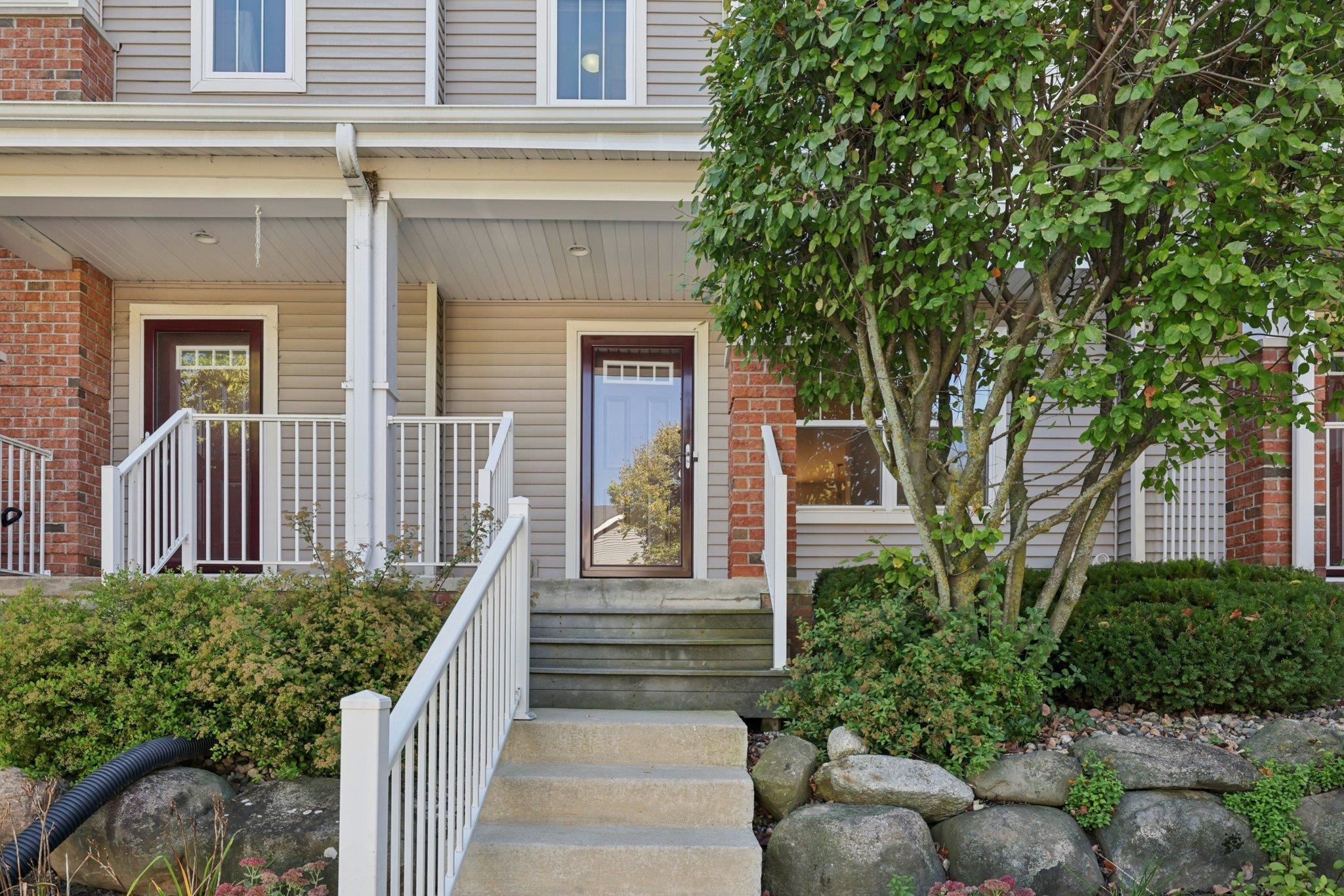Townhouse entrance with a covered porch, white railings, concrete steps, and a large leafy tree.