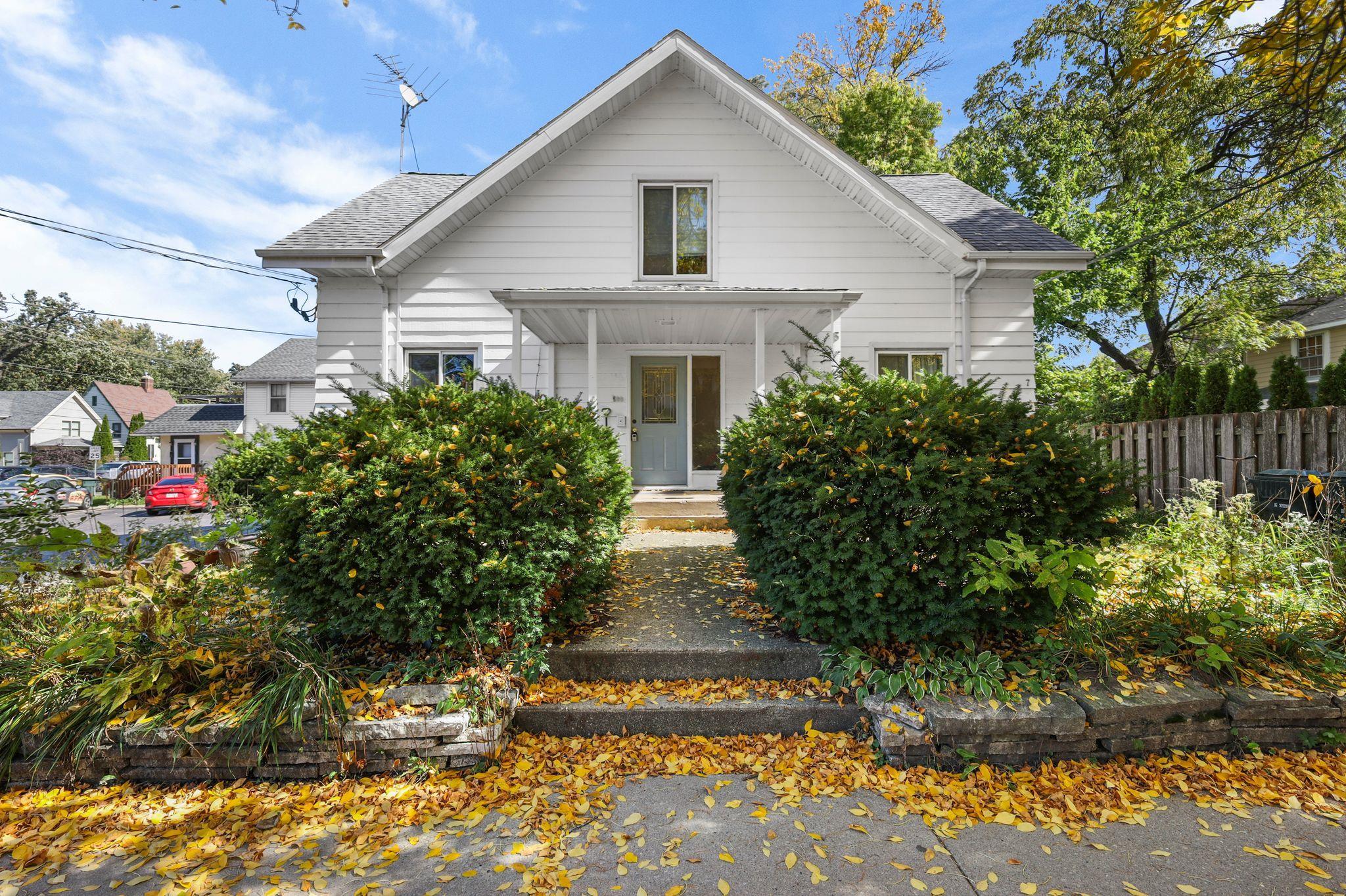 White gabled house with a covered porch, leafy shrubs, and autumn leaves along the front path.