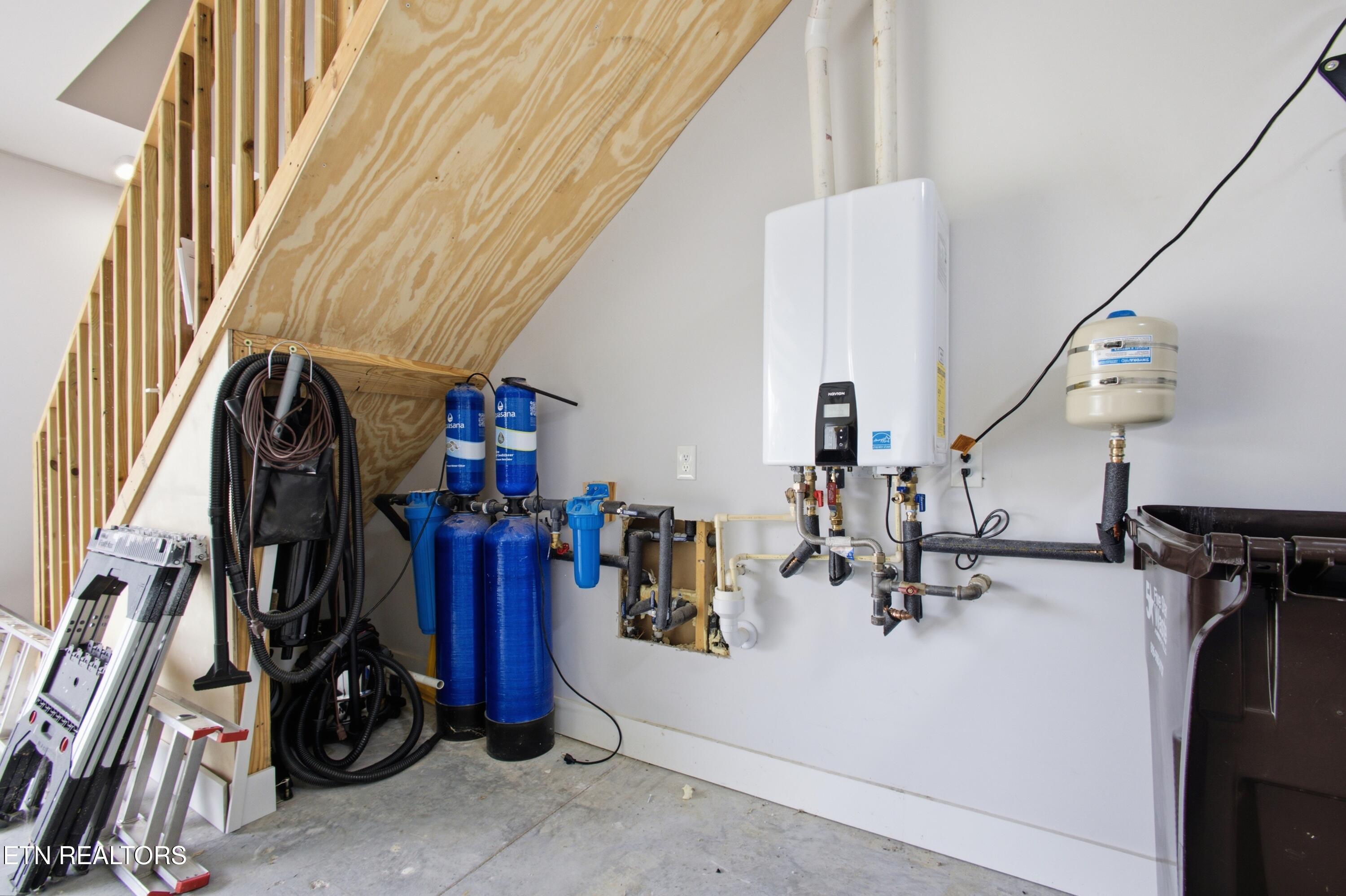 Utility area beneath an unfinished staircase with a tankless water heater and blue water filtration tanks.
