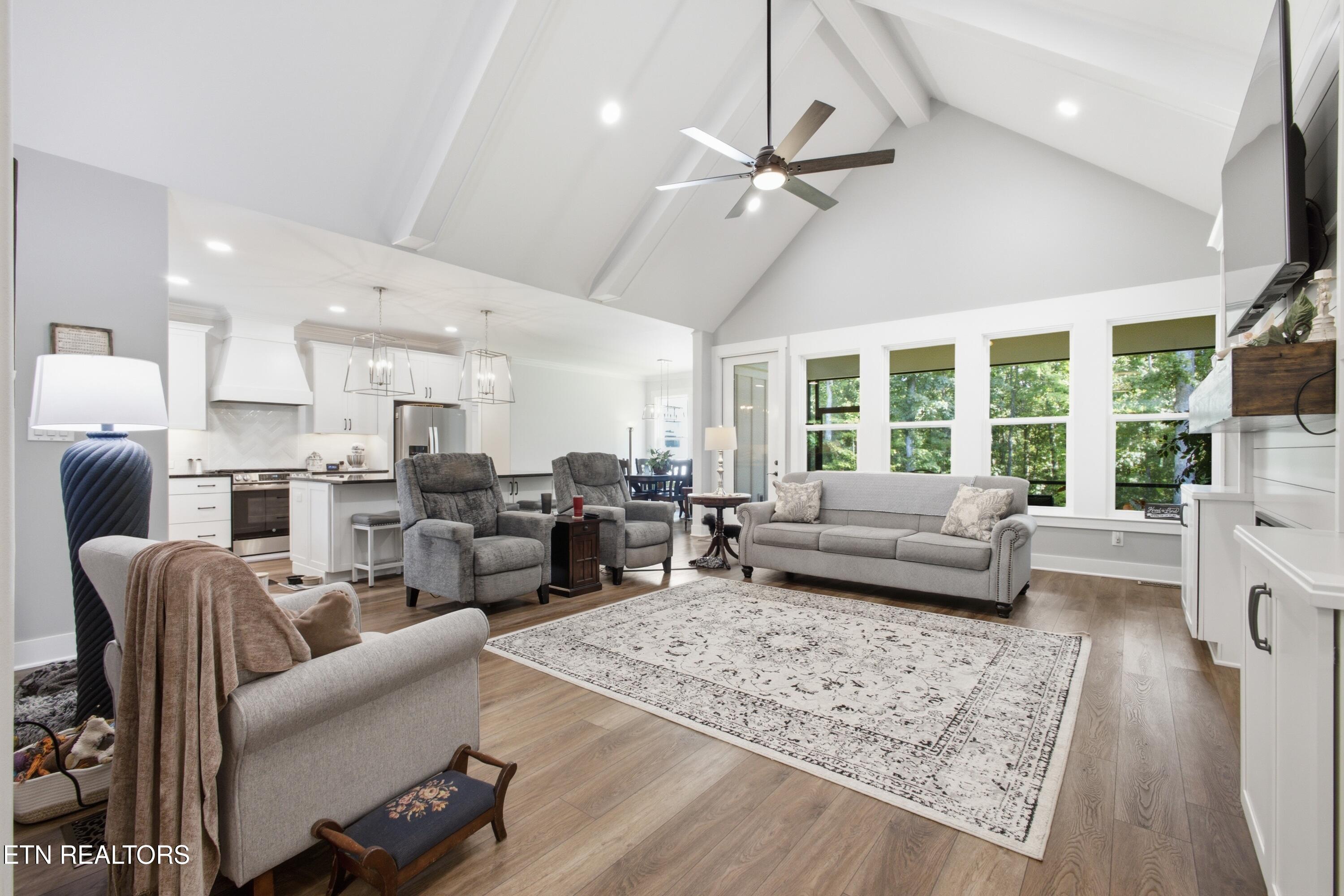 Open-plan living room and kitchen with vaulted ceiling, gray seating, wood floors, and large windows.
