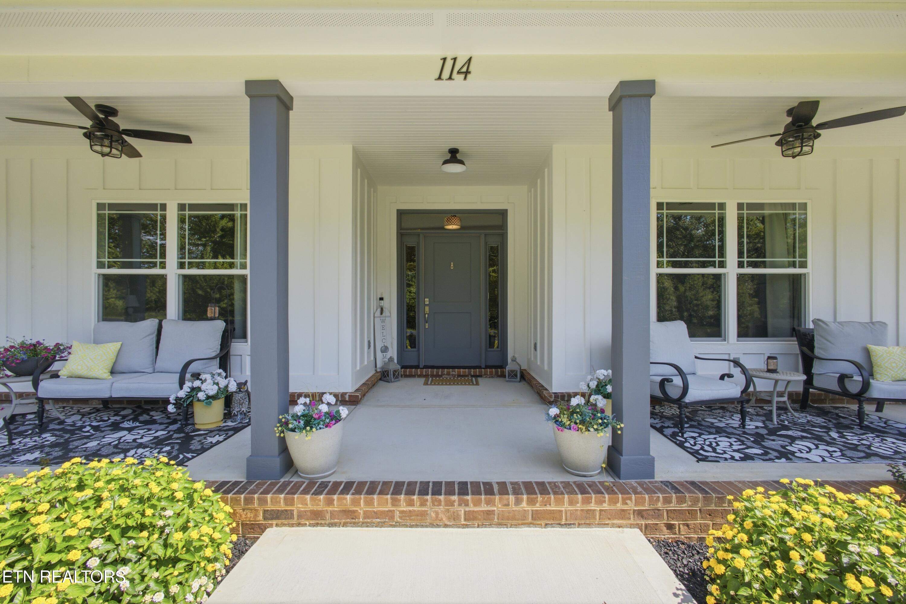 Home entrance with a covered, furnished porch, gray front door, brick steps, and potted flowers.