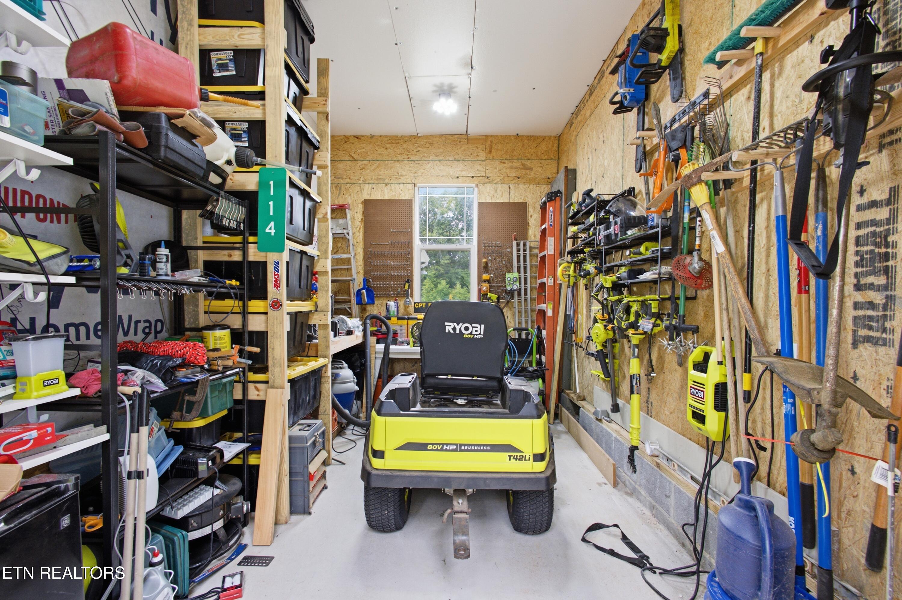 Garage workshop with a Ryobi riding mower, crowded storage shelves, and garden tools on the walls.