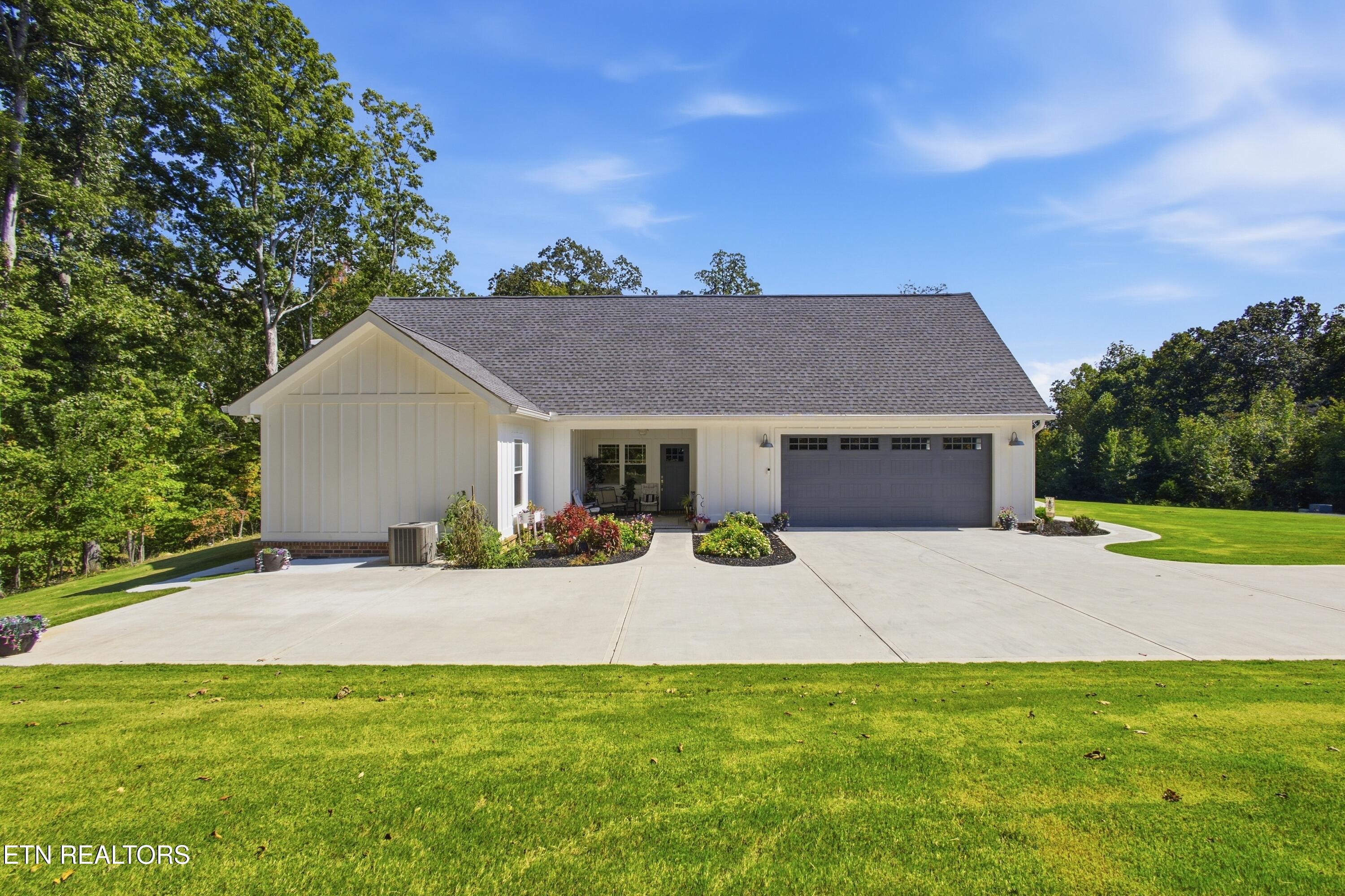 White home with a two-car garage, wide concrete driveway, landscaped entry, and lawn beside mature trees.