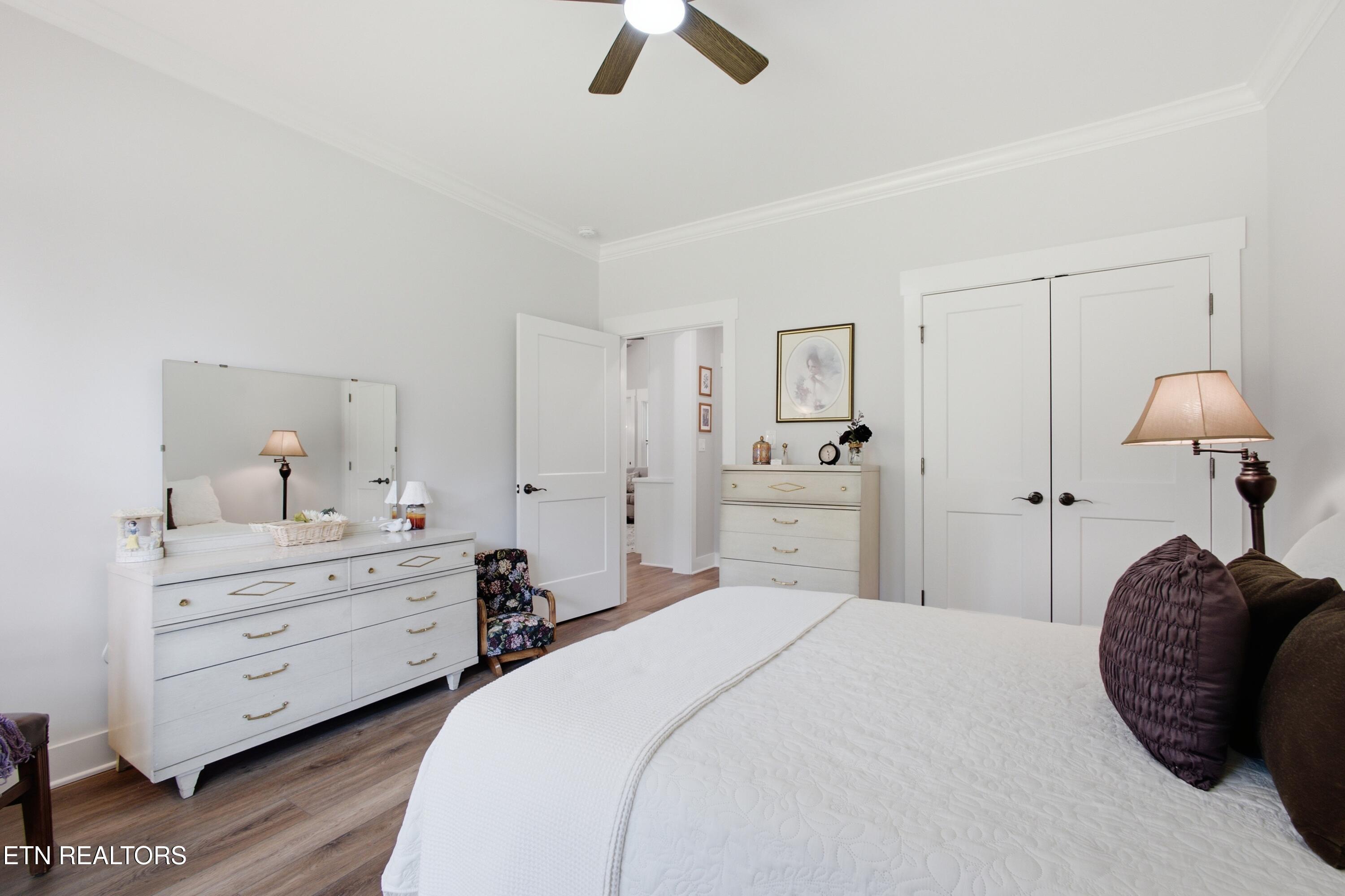 Bright bedroom with a large white bed, mirrored dresser, wood-look flooring, and double closet doors.
