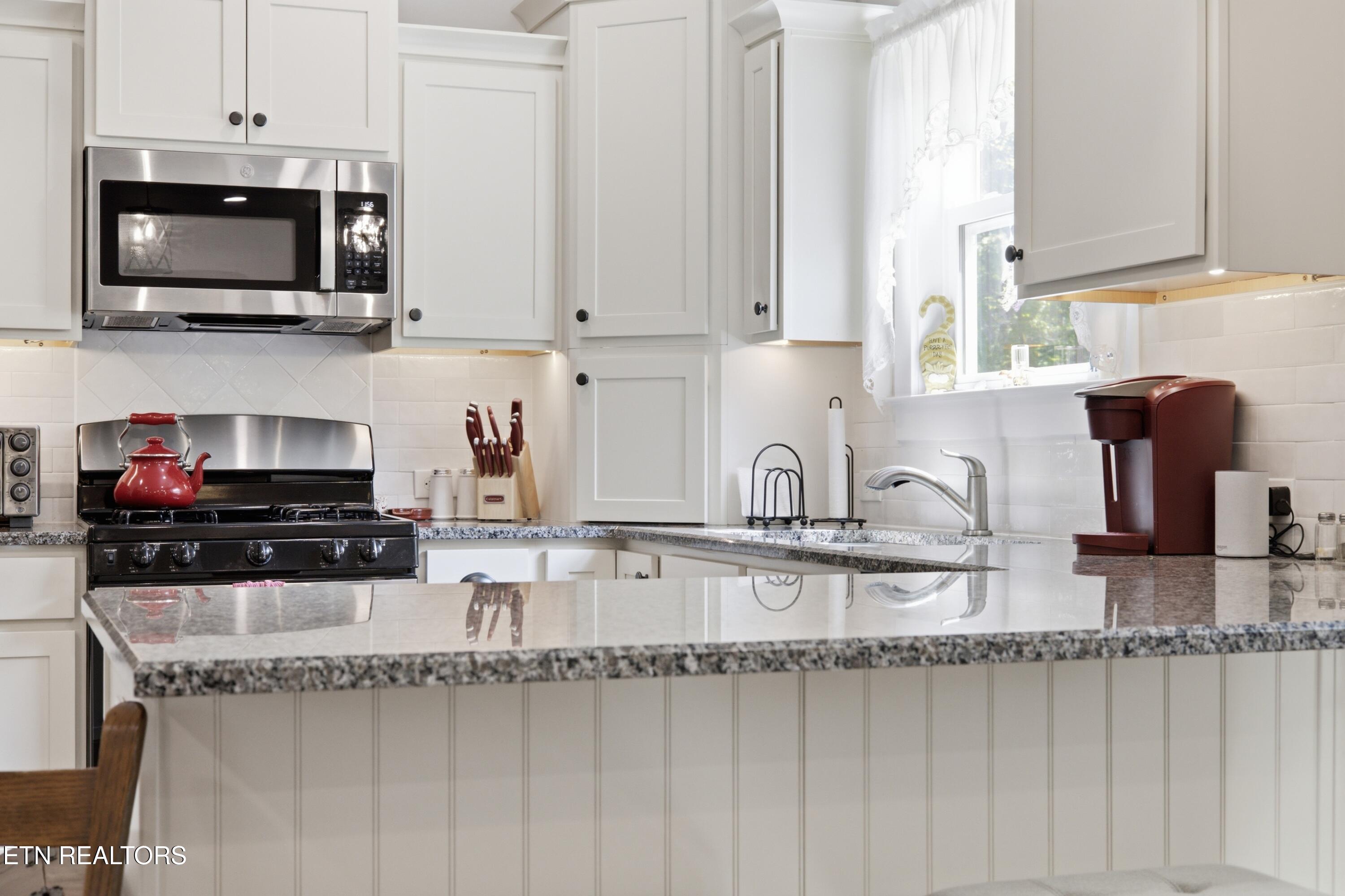 Kitchen with white cabinets, granite island, stainless microwave, and red kettle on the range.