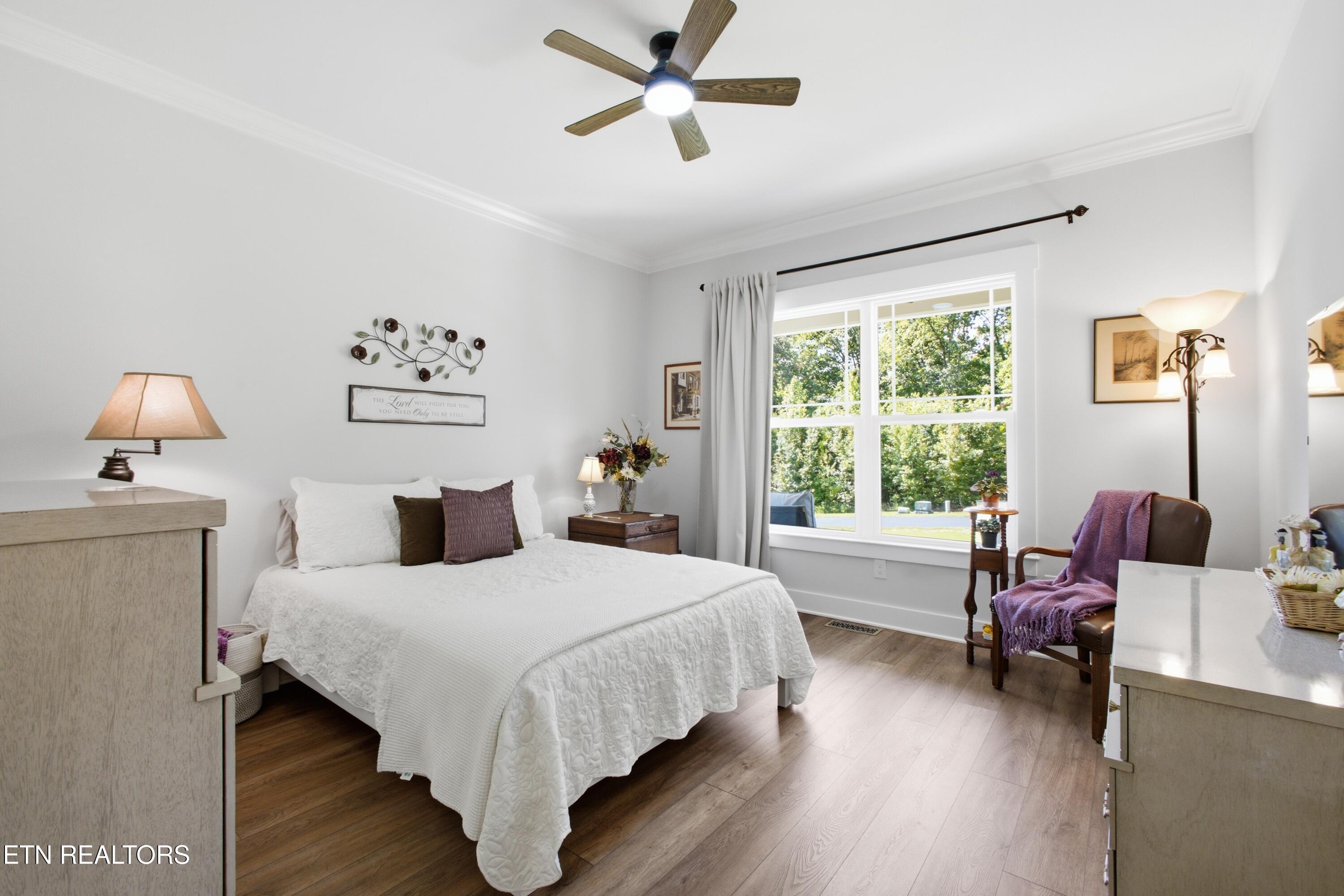 Furnished bedroom with a white quilted bed, wood flooring, and a large window overlooking trees.