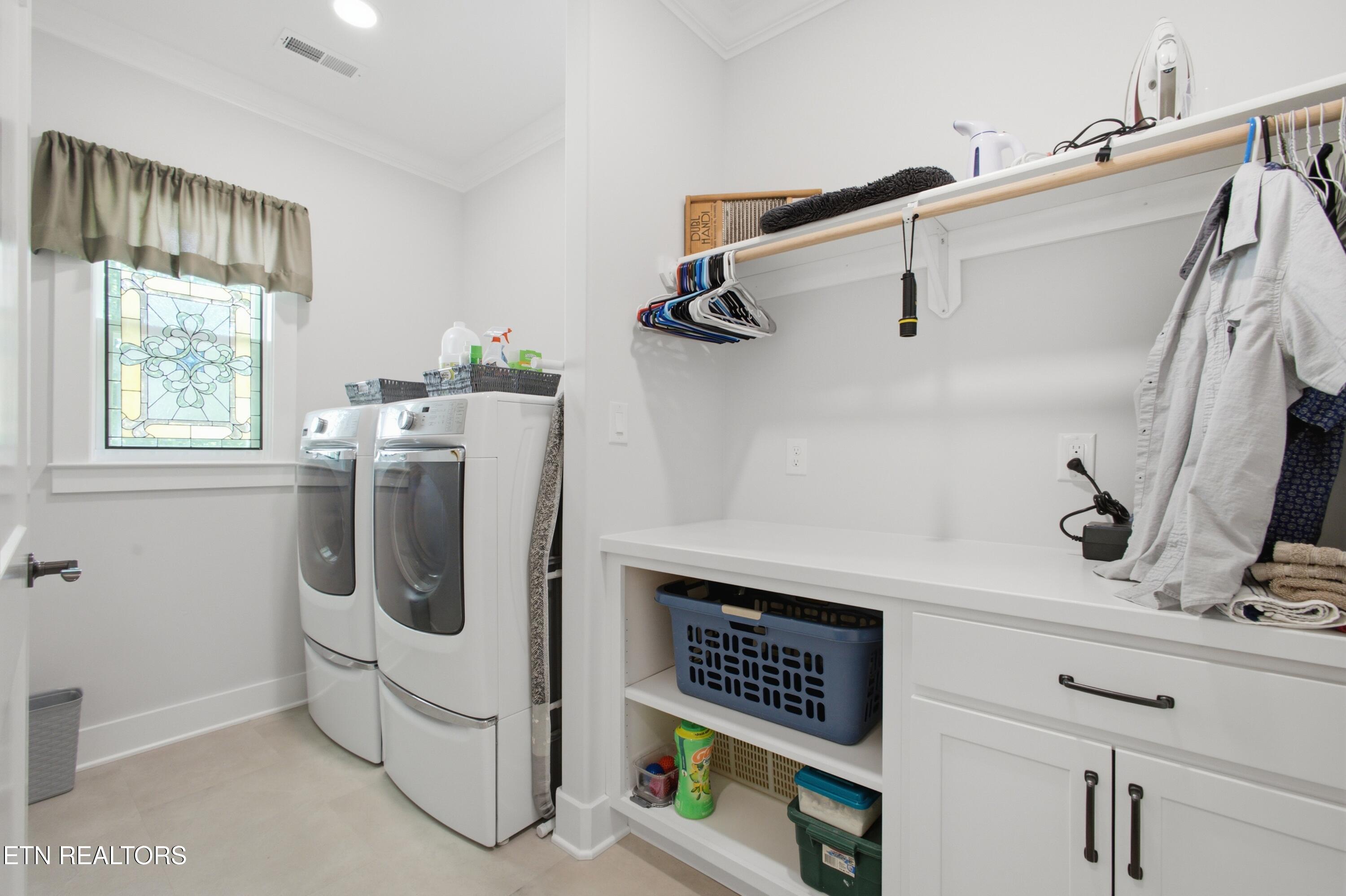 Laundry room with washer and dryer, white storage counter, hanging clothes, and stained-glass window.