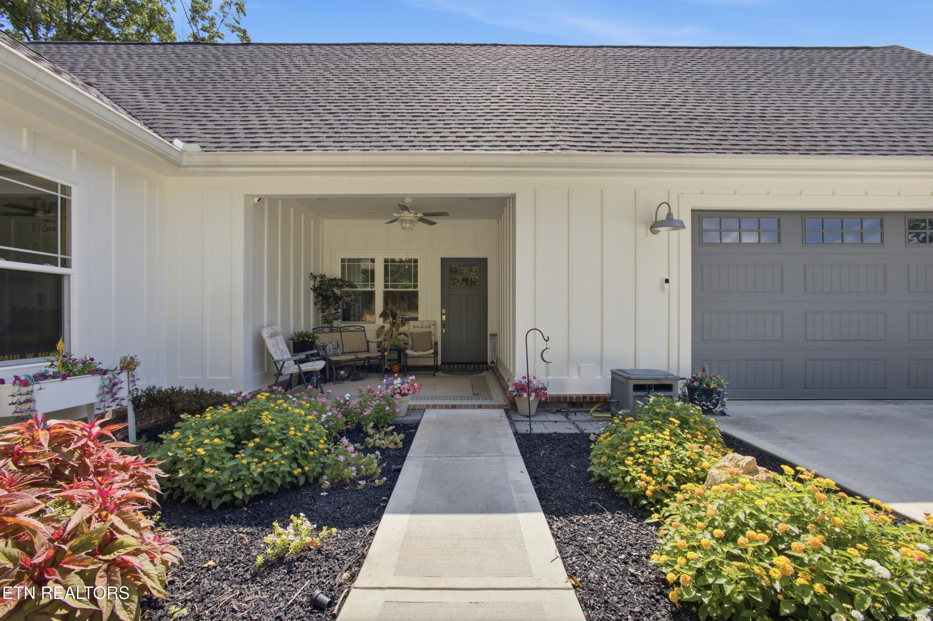 White home with a covered front porch, gray garage door, and colorful flower beds along the walkway.