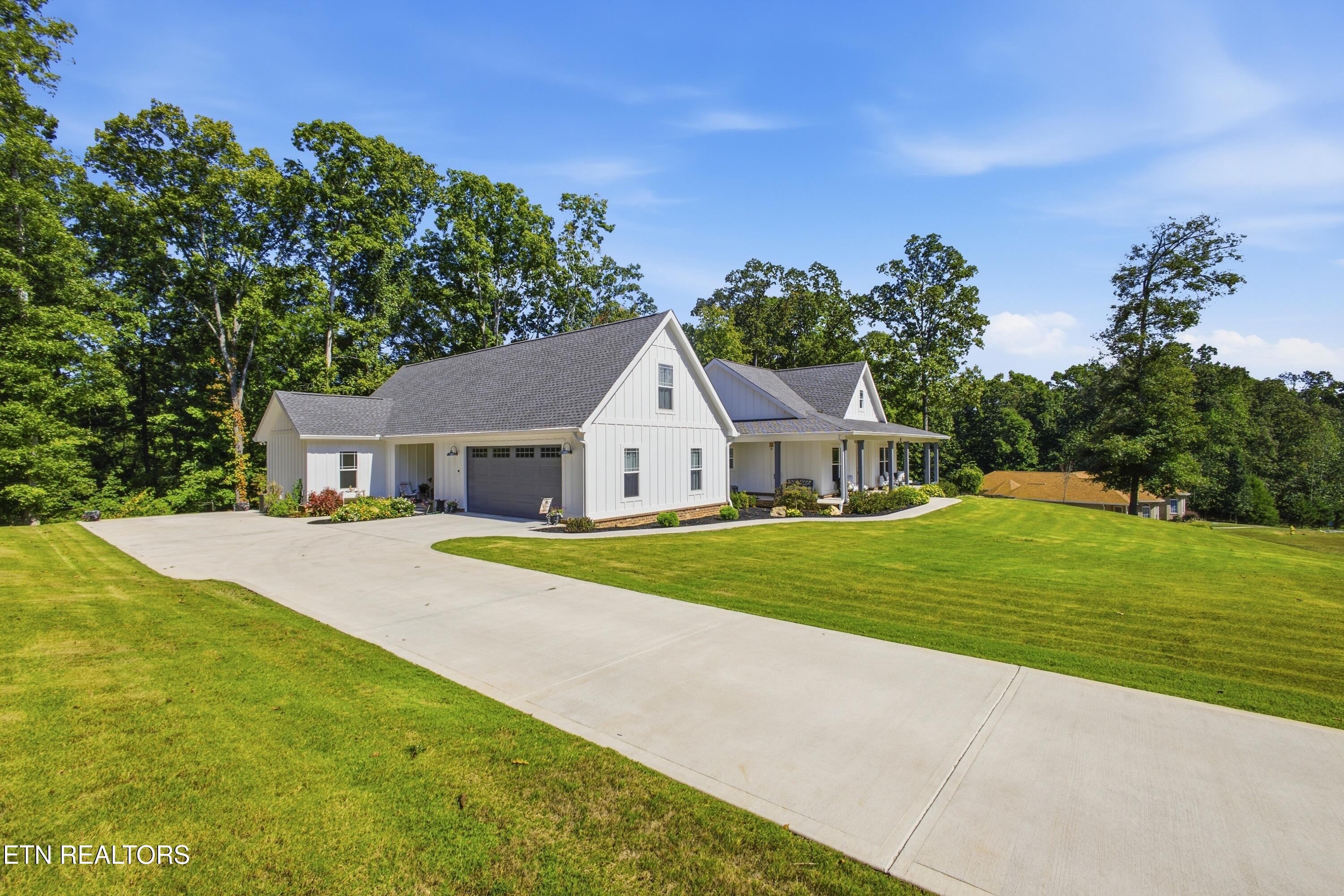 White gabled home with a front porch, attached garage, broad driveway, and mature trees.