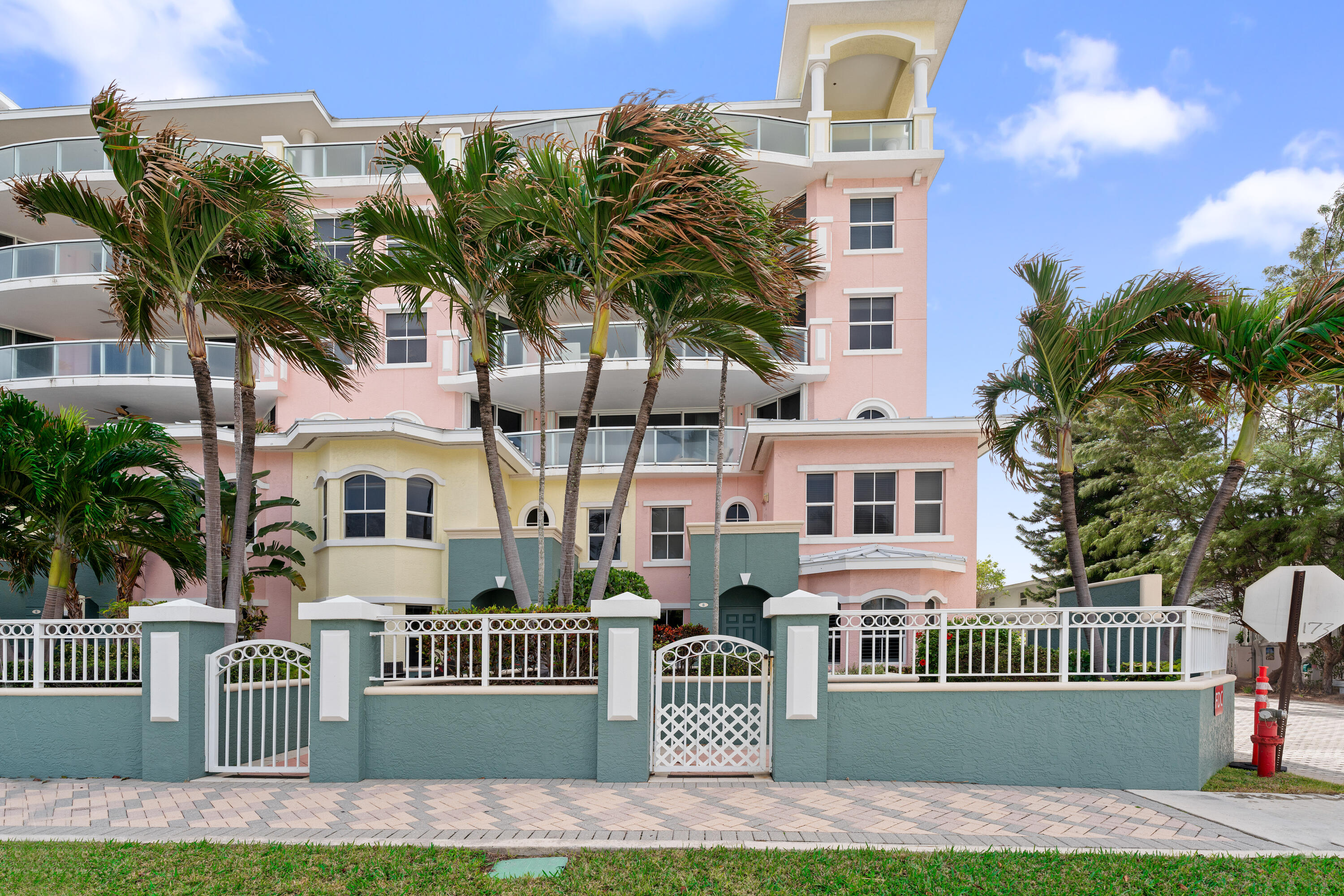 Pastel condominium exterior with glass balconies, palm trees, and a white gated entrance.