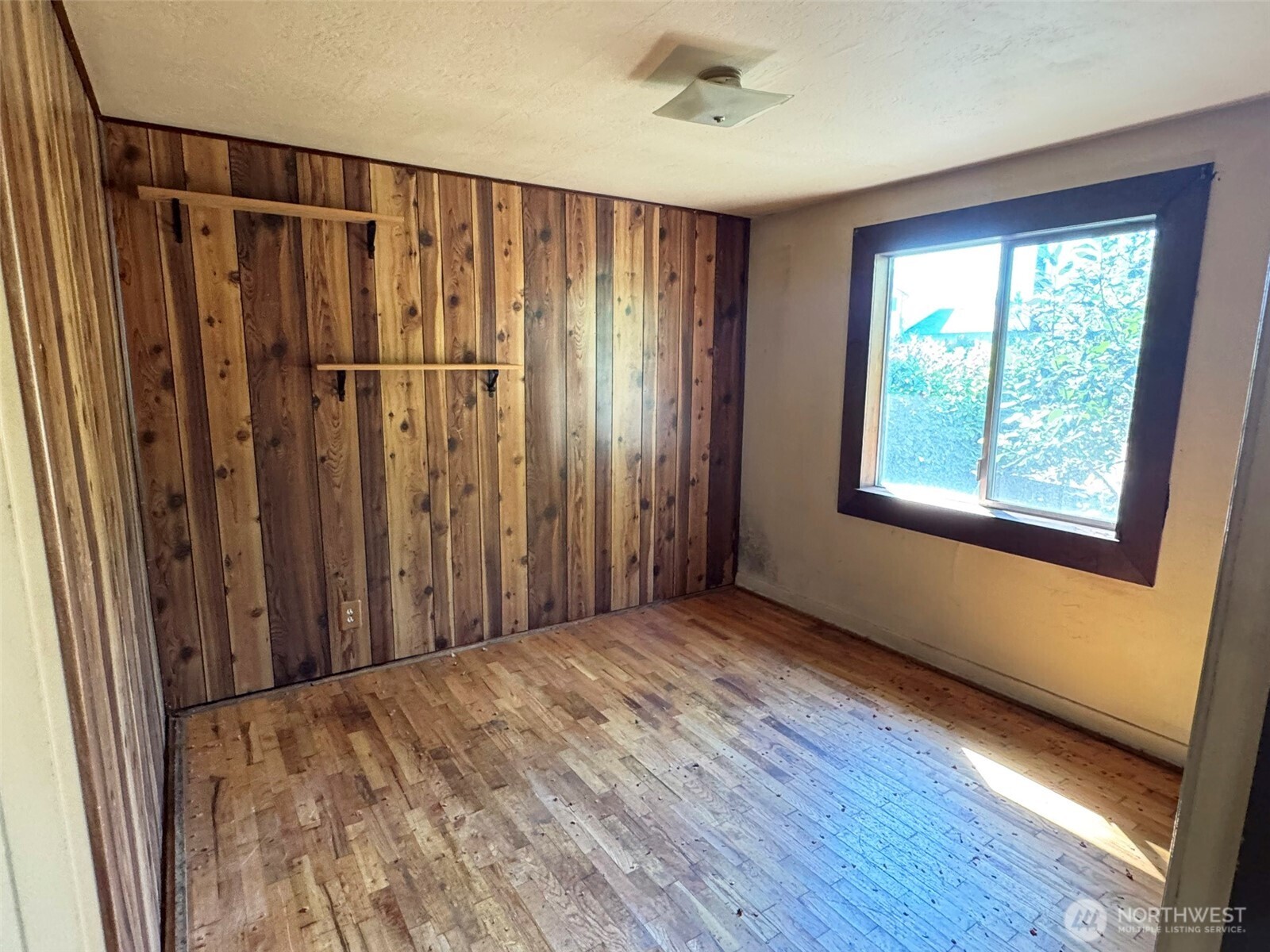 Empty room with wood-paneled walls, worn hardwood floors, and a large dark-trimmed window.