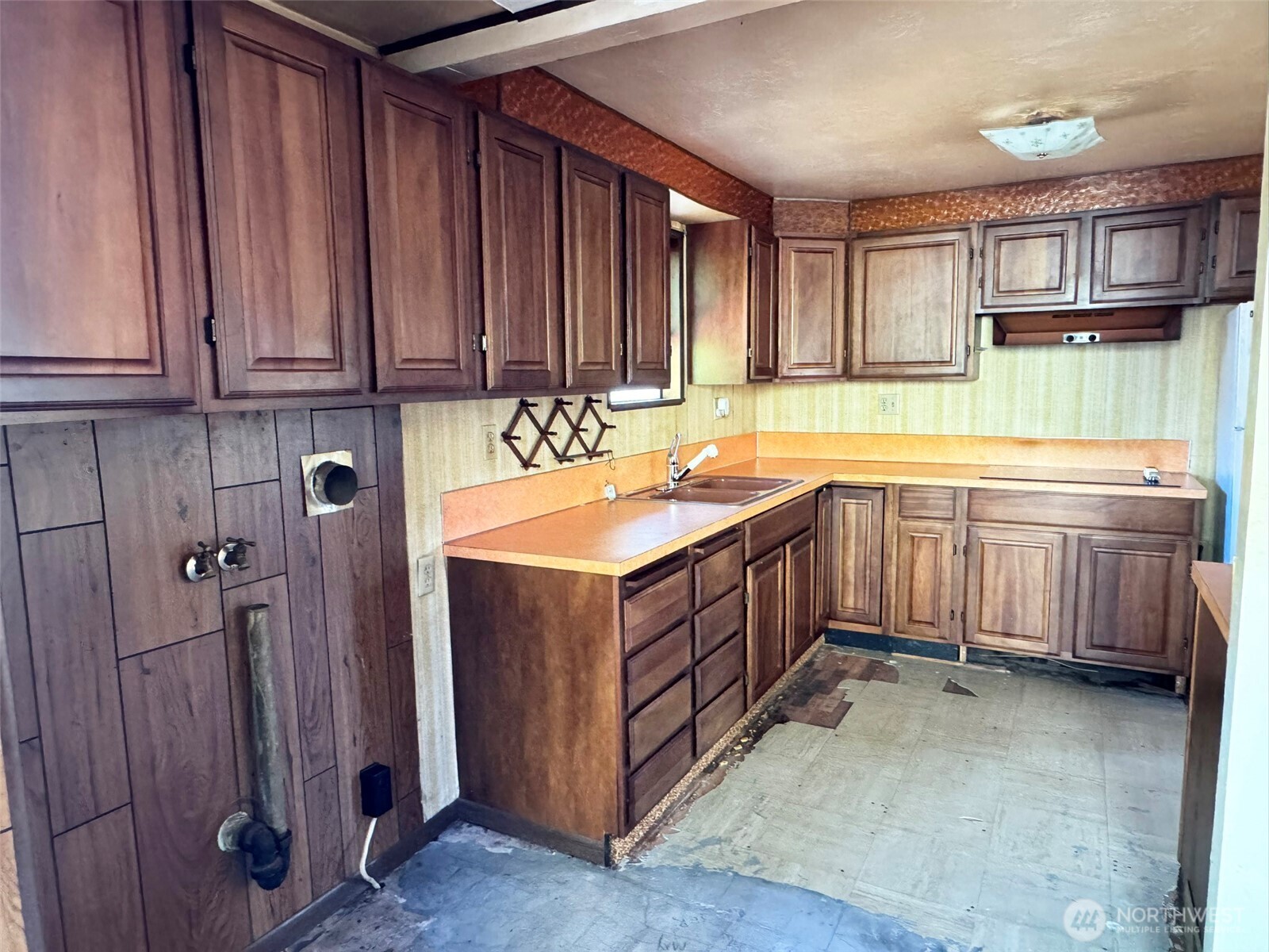 Empty kitchen with dark wood cabinets, orange counters, and worn unfinished flooring.
