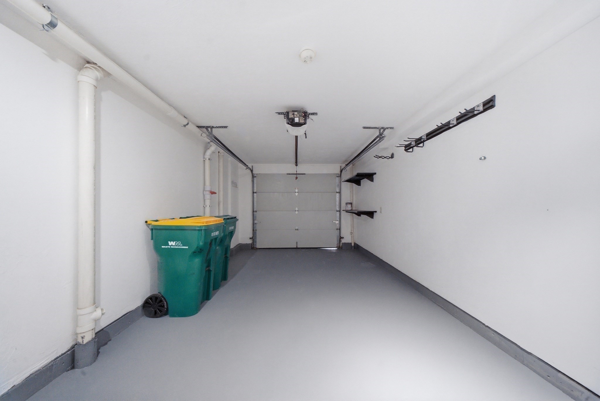 Empty garage with a gray floor, closed garage door, wall shelves, and green waste bins.
