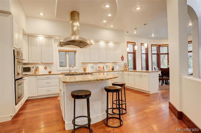 Kitchen with white cabinetry, stone island, stainless range hood, and warm wood floors.