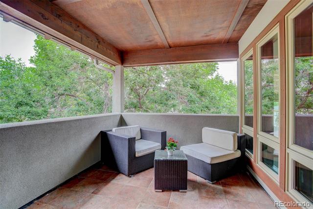 Covered patio with wicker seating, tiled floor, and large windows overlooking leafy trees.